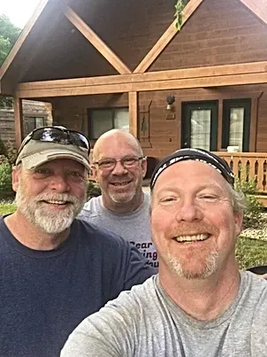 Three men are posing for a selfie in front of a log cabin.