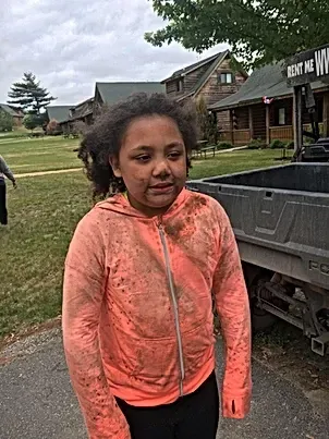 A young girl in a muddy jacket is standing in front of a truck.