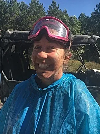 A woman wearing a blue poncho and goggles is smiling in front of a atv.