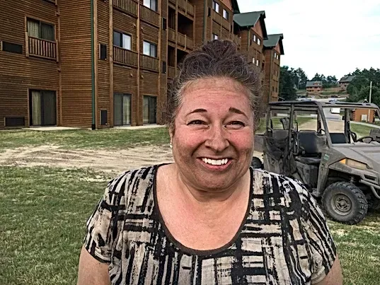 A woman is smiling in front of a building and a vehicle.