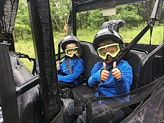 Two young boys wearing helmets and goggles are sitting in a vehicle giving a thumbs up.