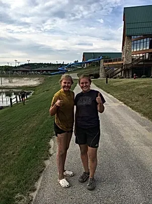 Two girls are standing next to each other on a sidewalk giving a thumbs up.
