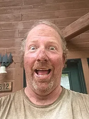 A man with a beard is making a funny face in front of a wooden wall.