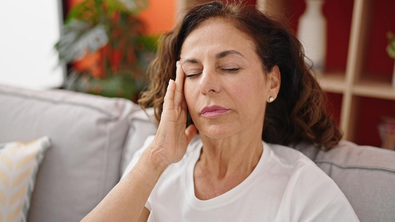 A woman is sitting on a couch with her eyes closed and holding her head.