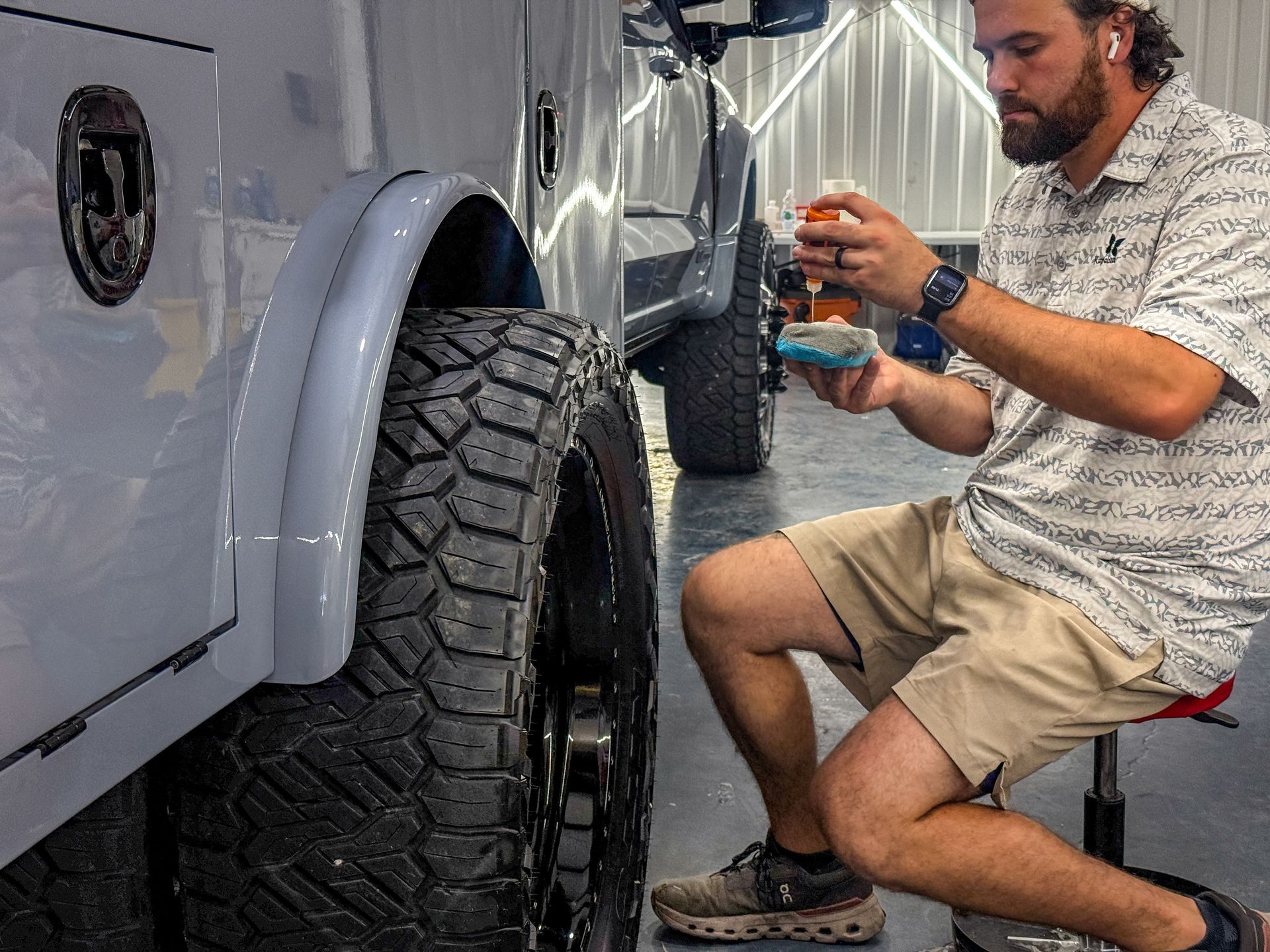Man cleans a truck tire with a brush and spray bottle indoors. He is wearing shorts and a patterned shirt.