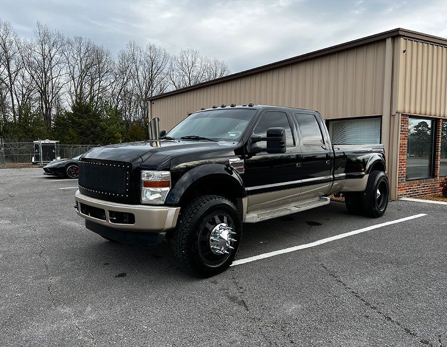Black and tan Ford F-350 dually truck parked in a paved lot, near a tan building with a cloudy sky.