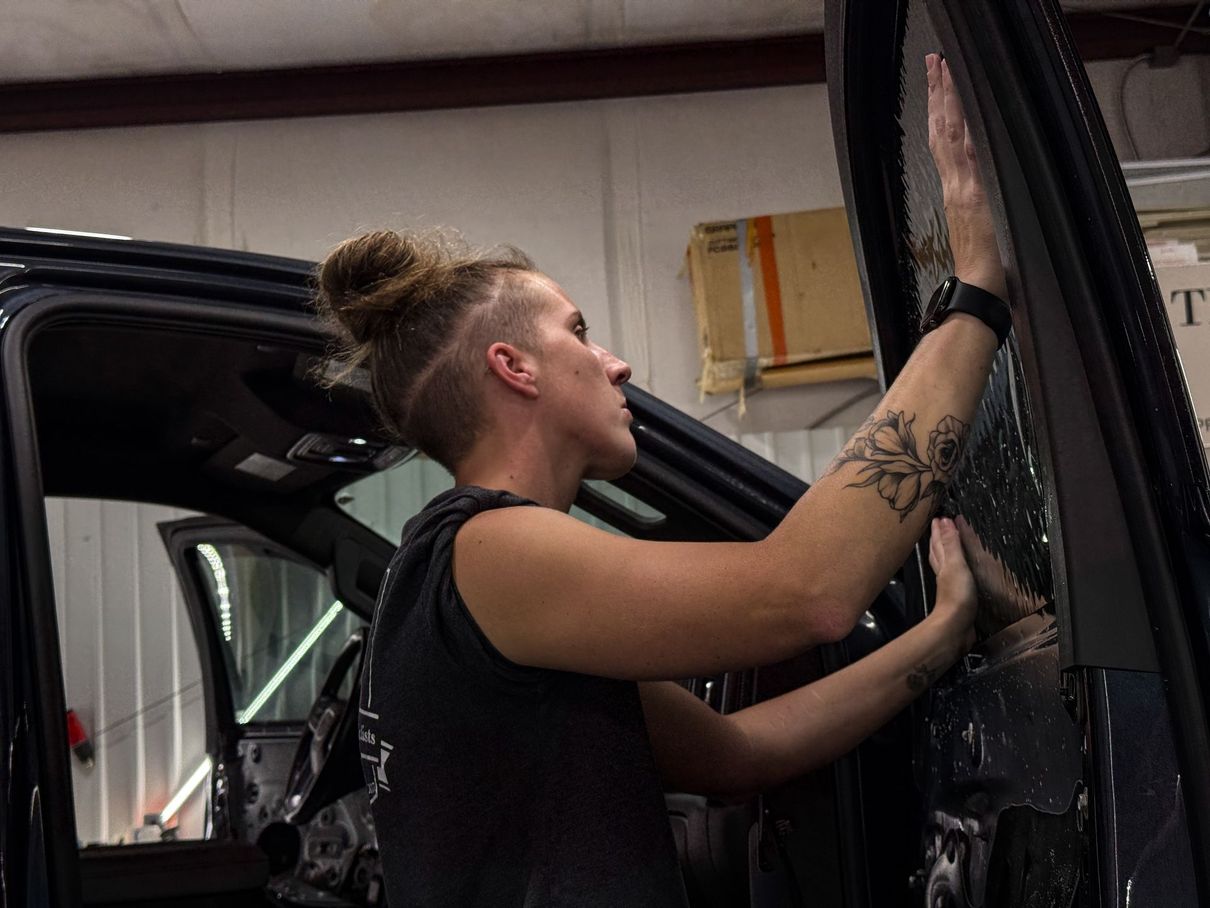 A person with a bun and tattoos applying tint to a car window inside a garage.
