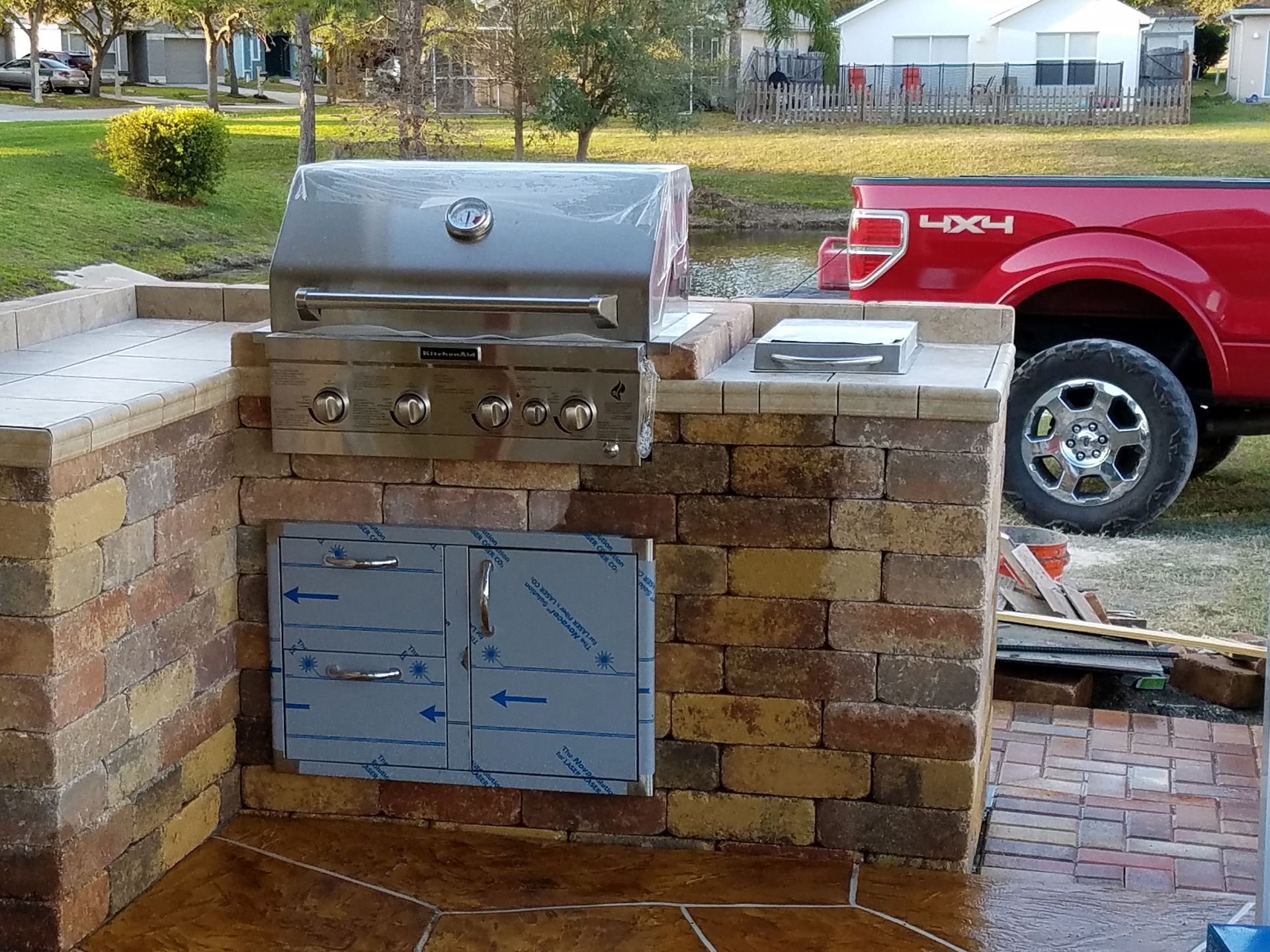 Outdoor brick grilling station with a stainless steel grill, cabinet doors, and a red pickup truck in the background.