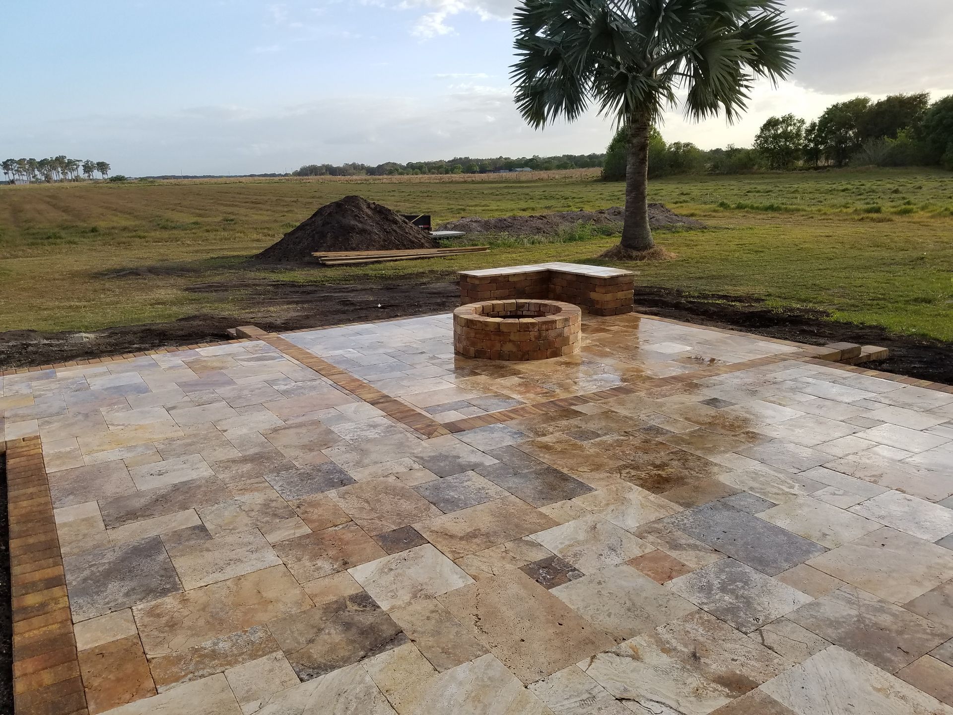 Finished stone patio with fire pit in a field, a small palm tree in the background.