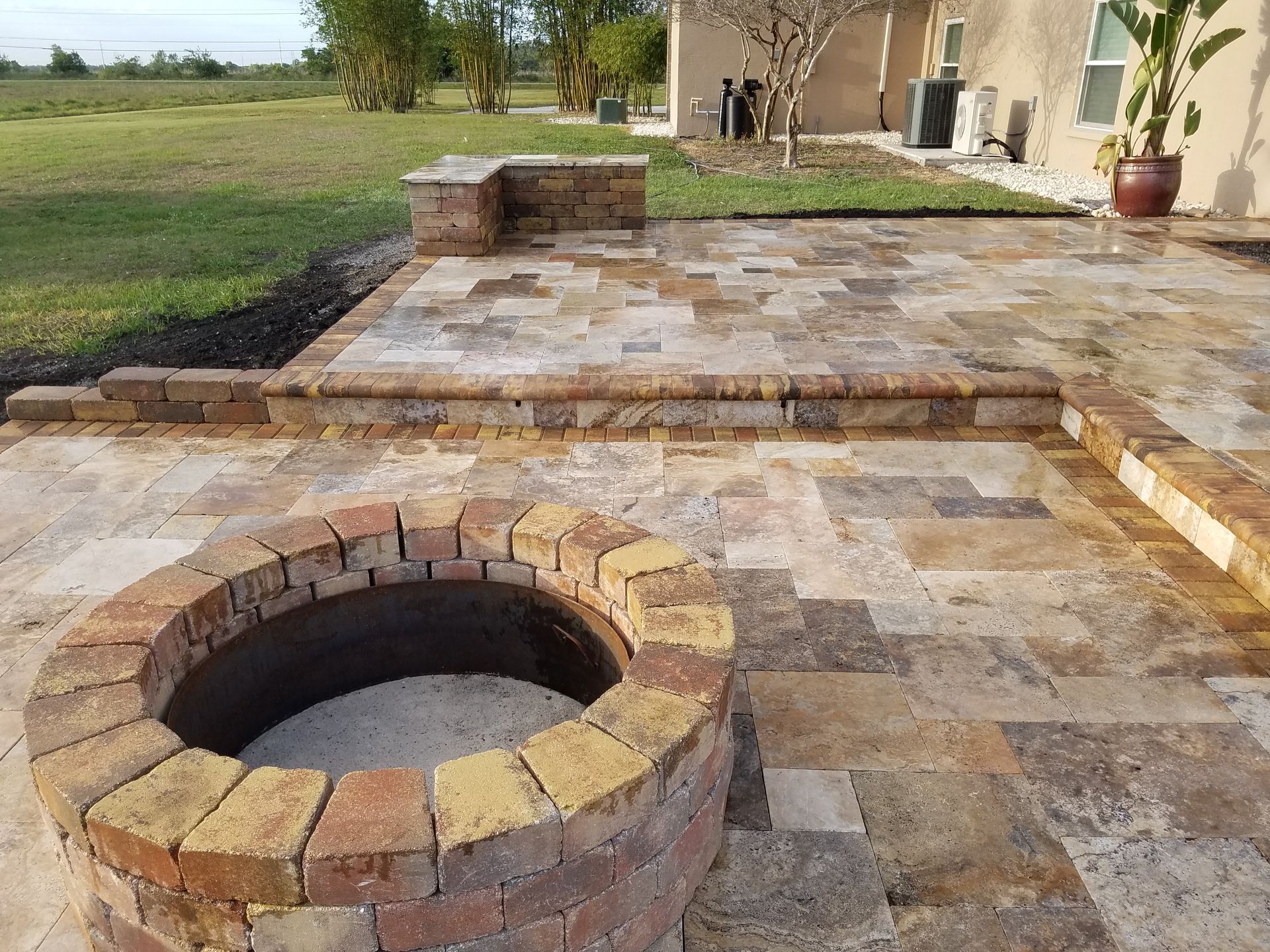 Brick patio with a circular fire pit, low wall, and steps. The area includes tan paving stones and a grassy background.