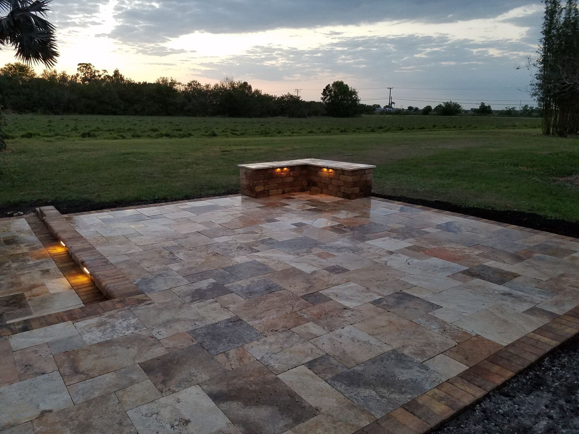 Stone patio with a built-in fire pit, overlooking a grassy field under a twilight sky.