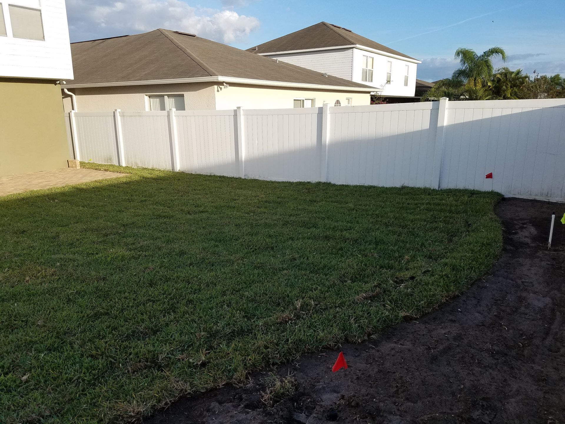 Green lawn in a backyard bordered by a white fence and dirt. Two houses are visible in the background under a blue sky.