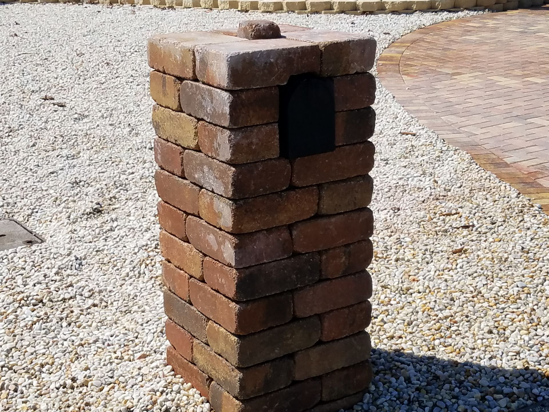 A brick pillar with a rectangular opening, sitting on gravel. It's a brown and tan color and outdoors.
