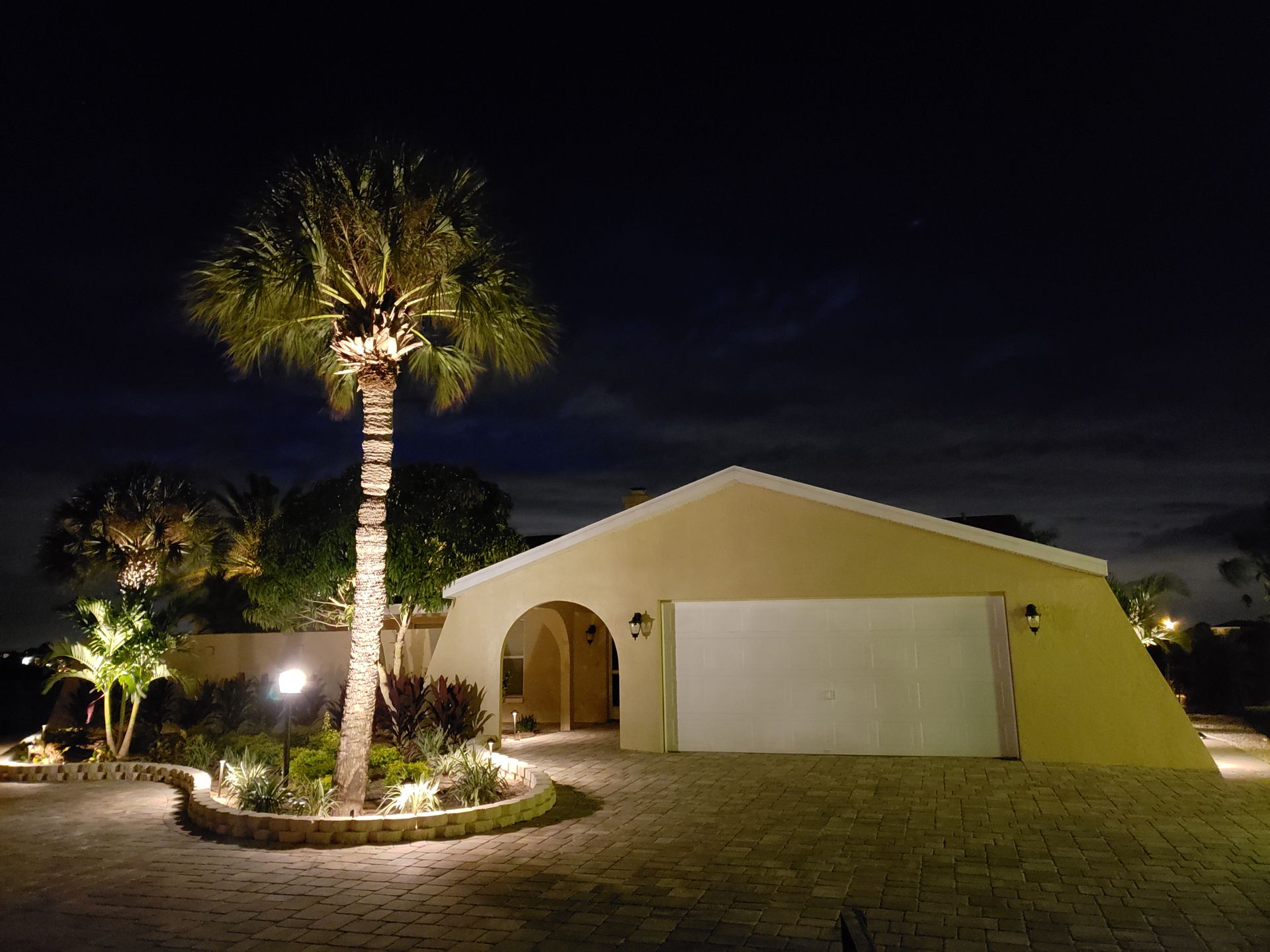 A house at night with illuminated landscaping. A palm tree, driveway, and garage are lit by spotlights.