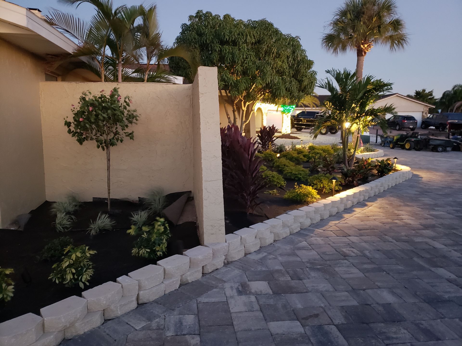 A well-lit, landscaped driveway and garden bed at dusk, with stacked stone border and colorful plants against a beige wall.