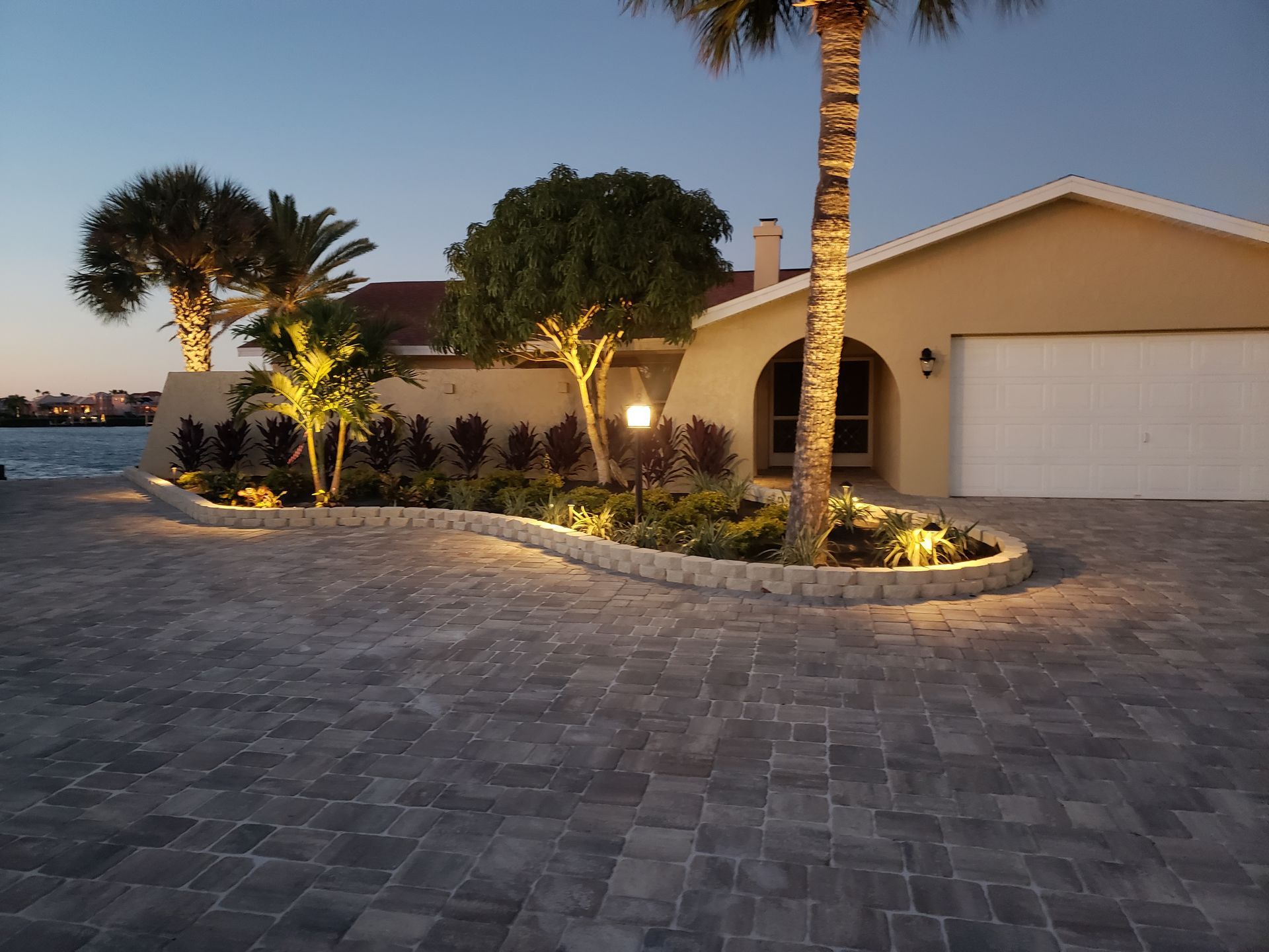 A house with a paved driveway and landscaping illuminated by lights at dusk. Palm trees and other plants are visible.