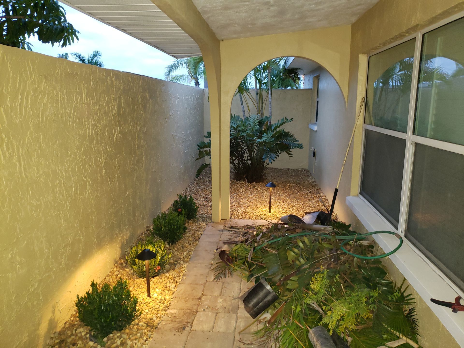 A narrow outdoor walkway with beige walls, small bushes, and a stone path leads to a small courtyard with greenery.