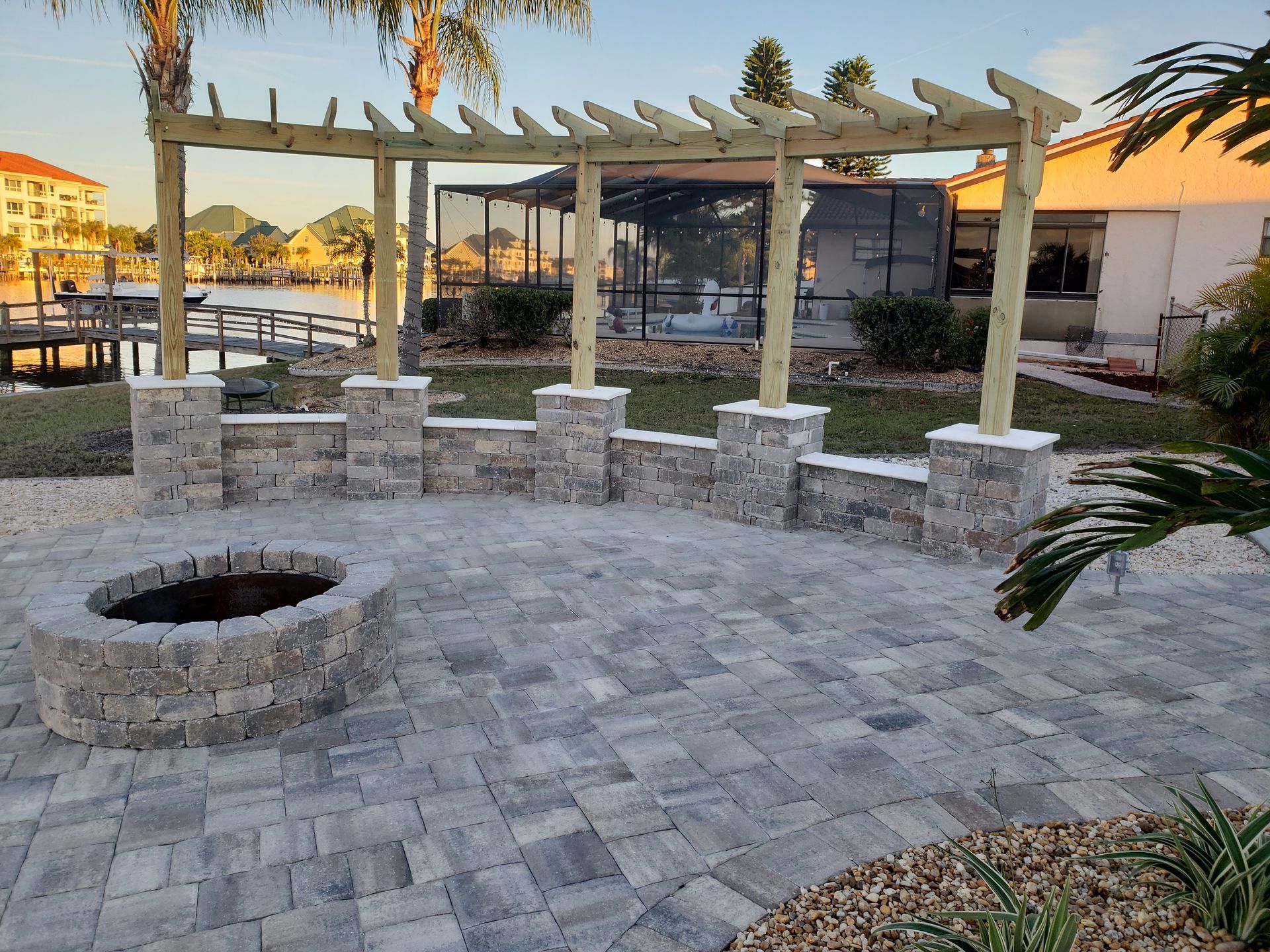 A backyard patio with a fire pit, brick wall, pergola, and water view. Blue-gray pavers cover the ground.
