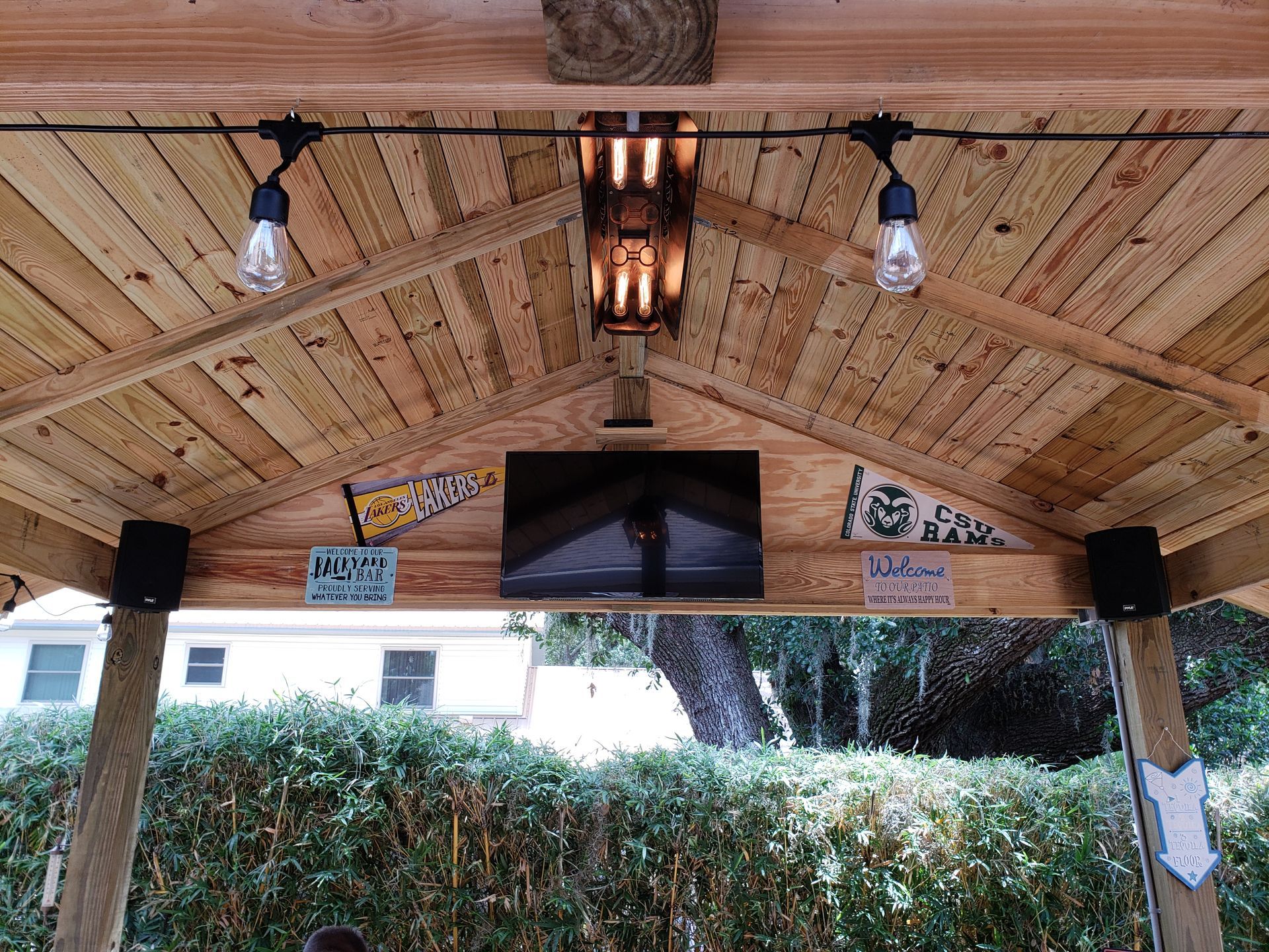 Wooden outdoor structure with string lights, a mounted TV, and speakers, set against a background of trees and a building.