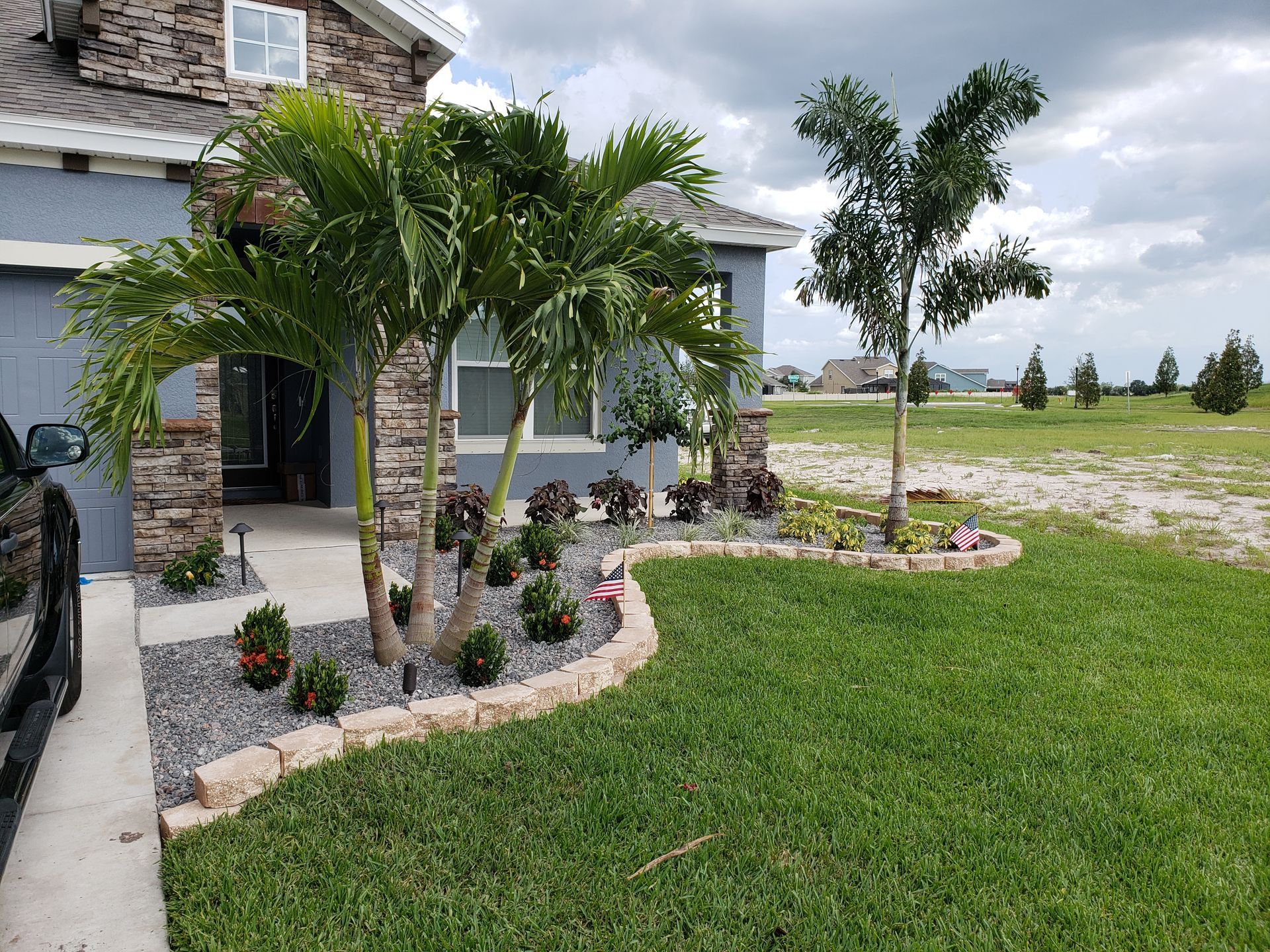 Front yard landscaping with a house featuring stone accents and a palm tree. A lush green lawn and a cloudy sky are visible.