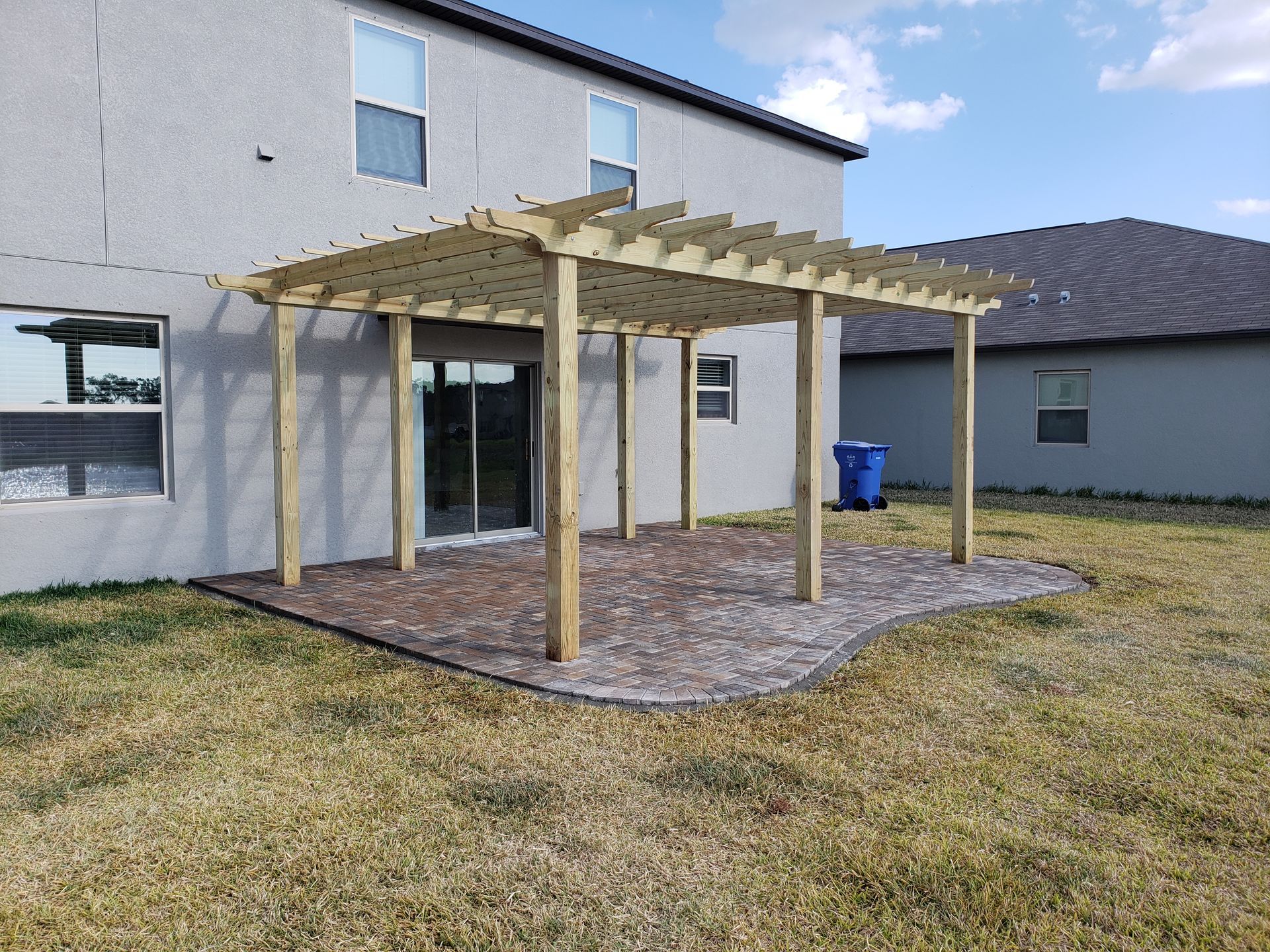 Pergola structure attached to a light gray house, over a patio area. A blue trash can sits in the background.