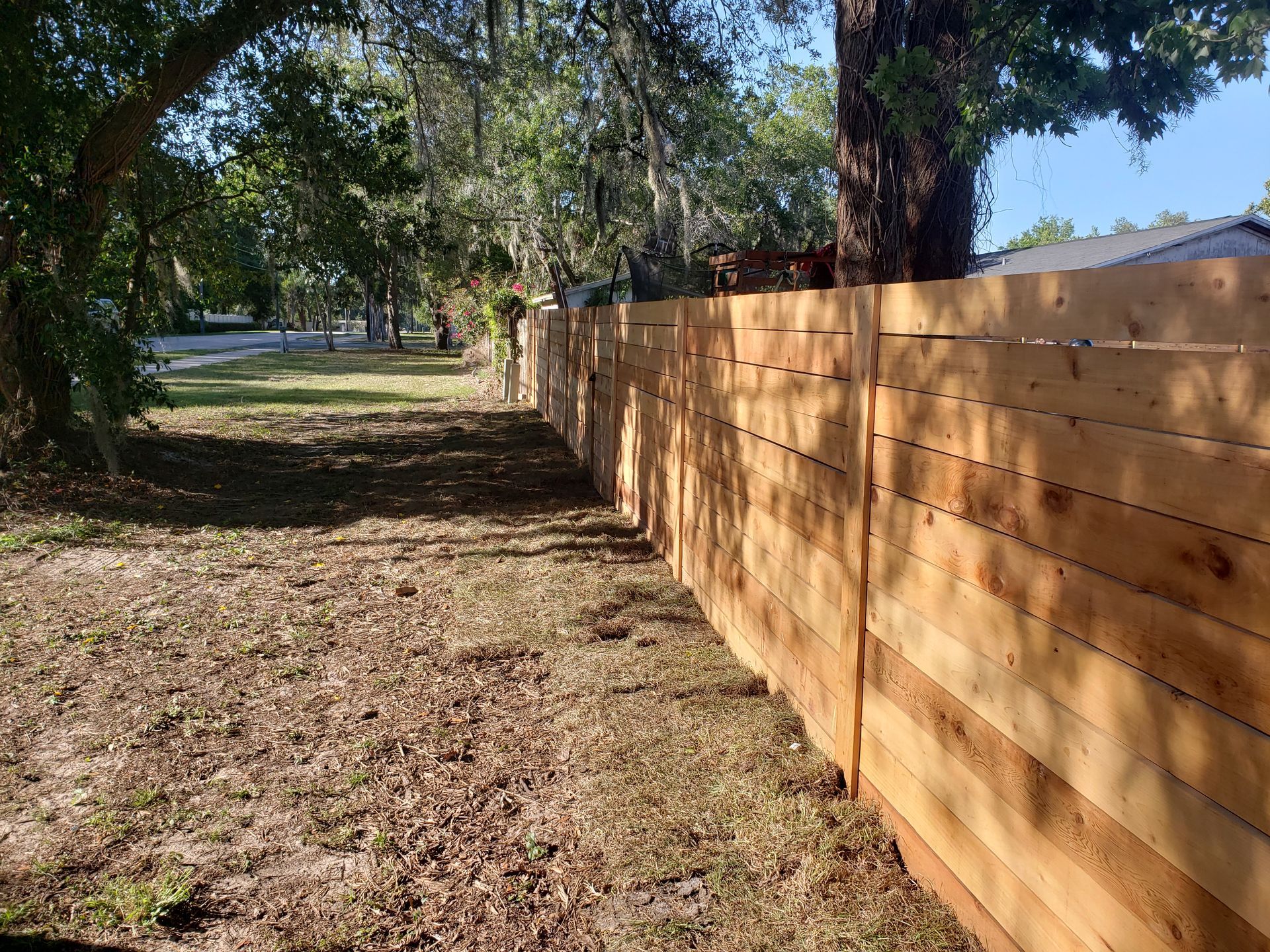A horizontal wooden fence runs along a grassy yard, with trees in the background. The fence is brown.