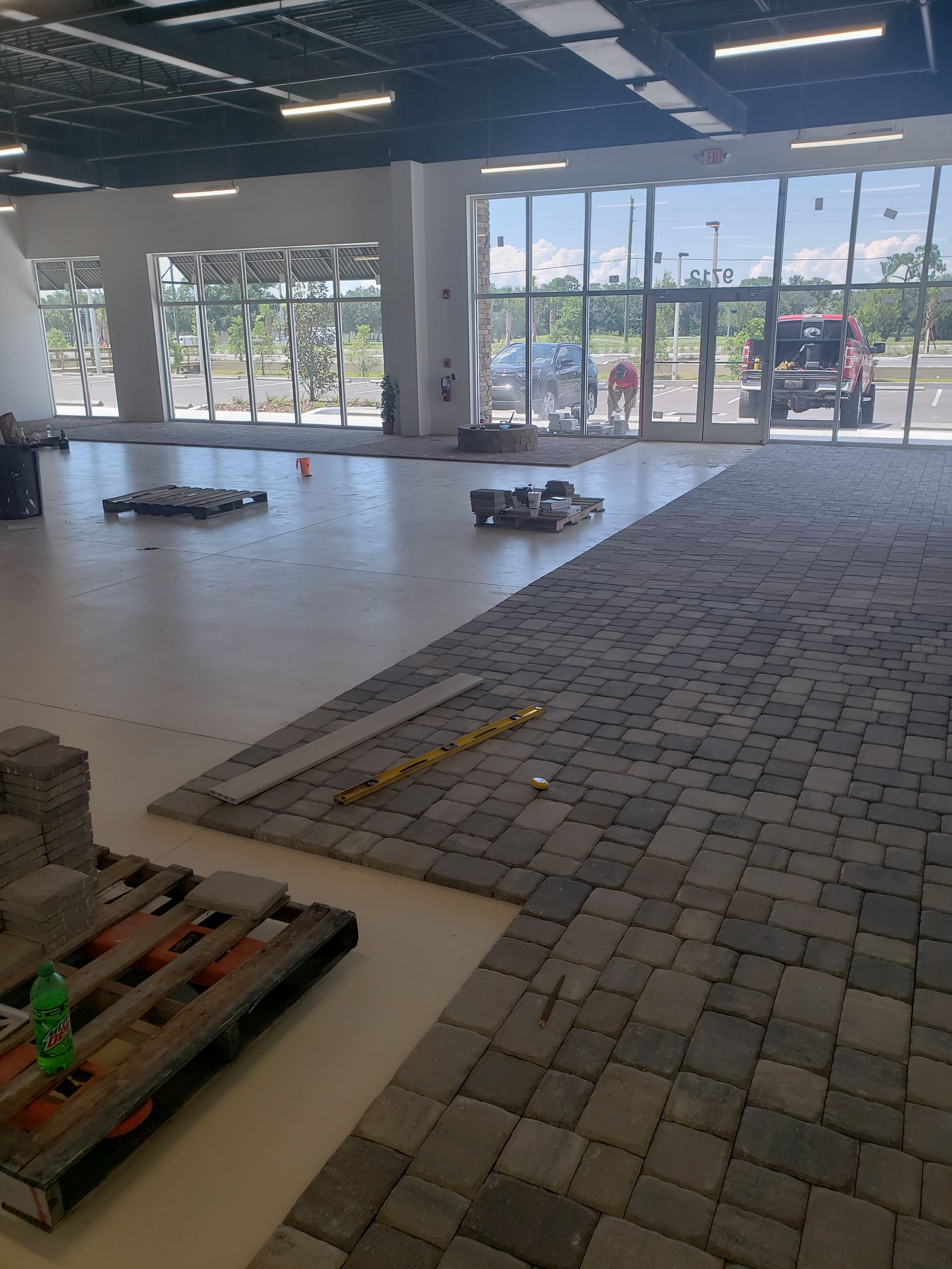 Interior space with workers installing gray brick flooring. Pallets and tools scattered about, large windows in the background.