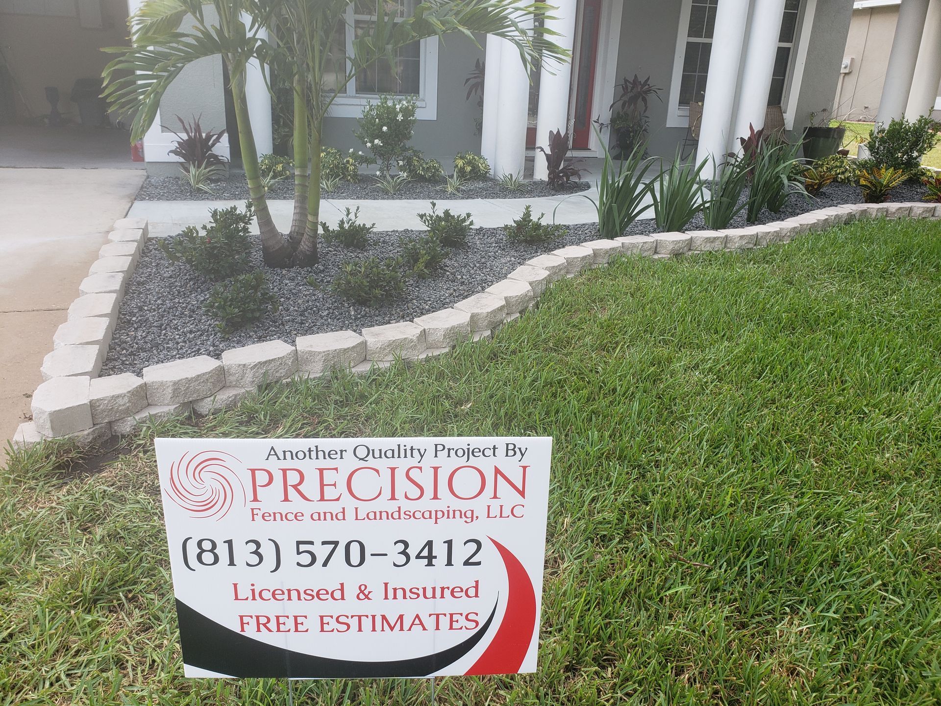 A sign for Precision Fence and Landscaping in front of a house with a landscaped yard and a stone border.