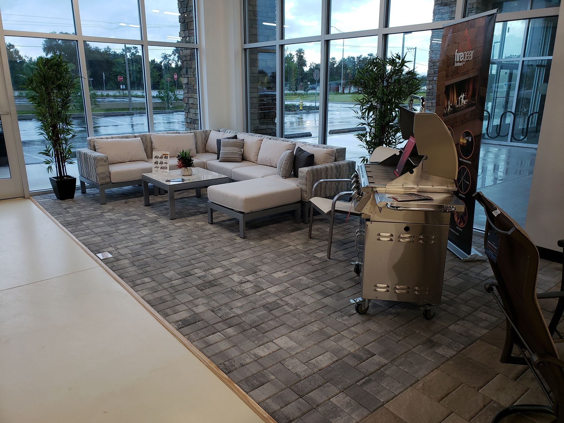 Indoor showroom with outdoor furniture. Light-colored sectional sofa, grill, and artificial plants sit on a gray brick-patterned rug. Large windows show an overcast sky.