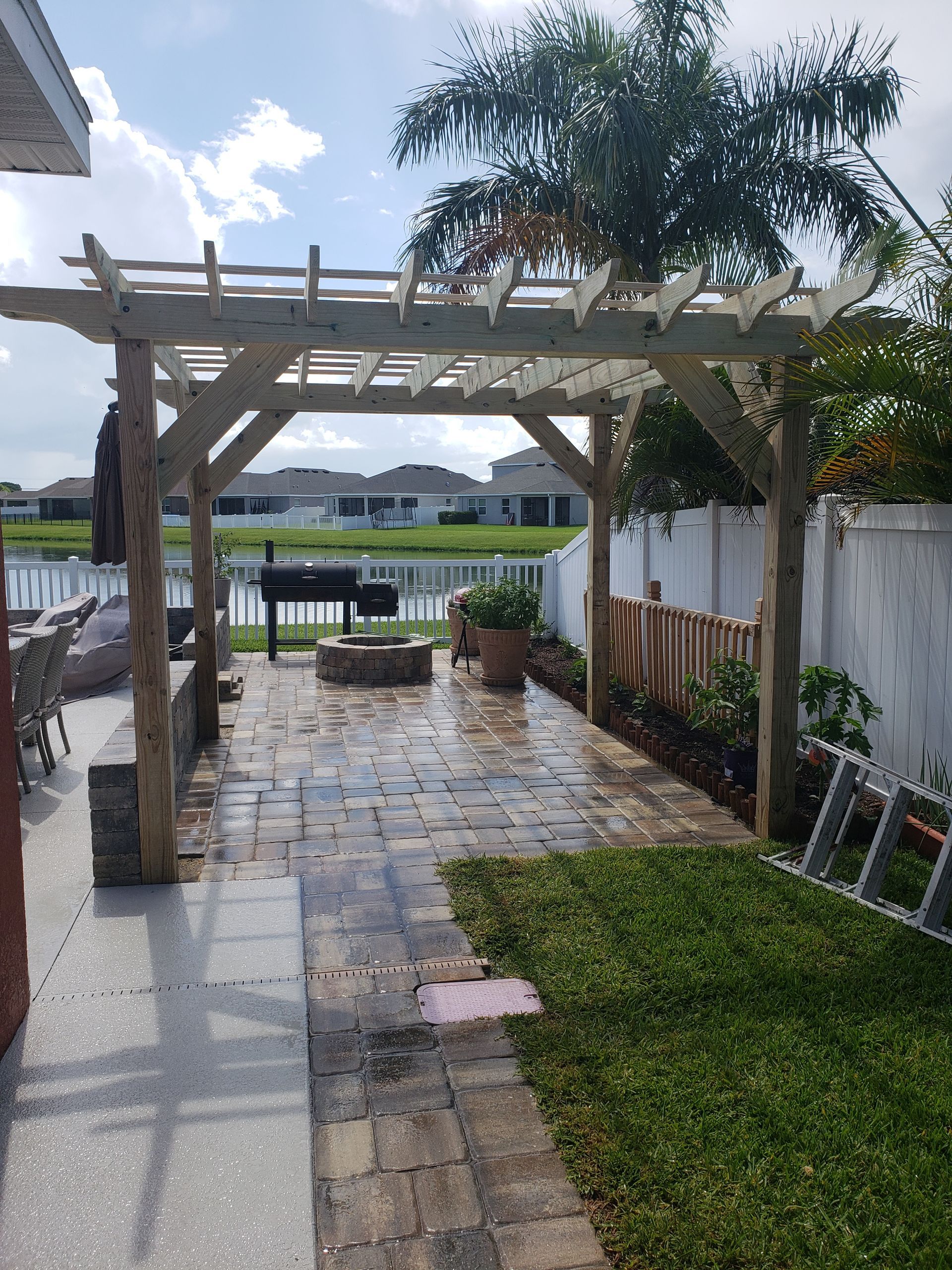 A backyard patio with a pergola, brick pavers, a grill, fire pit, and lush green grass. A white fence and water are visible in the background.