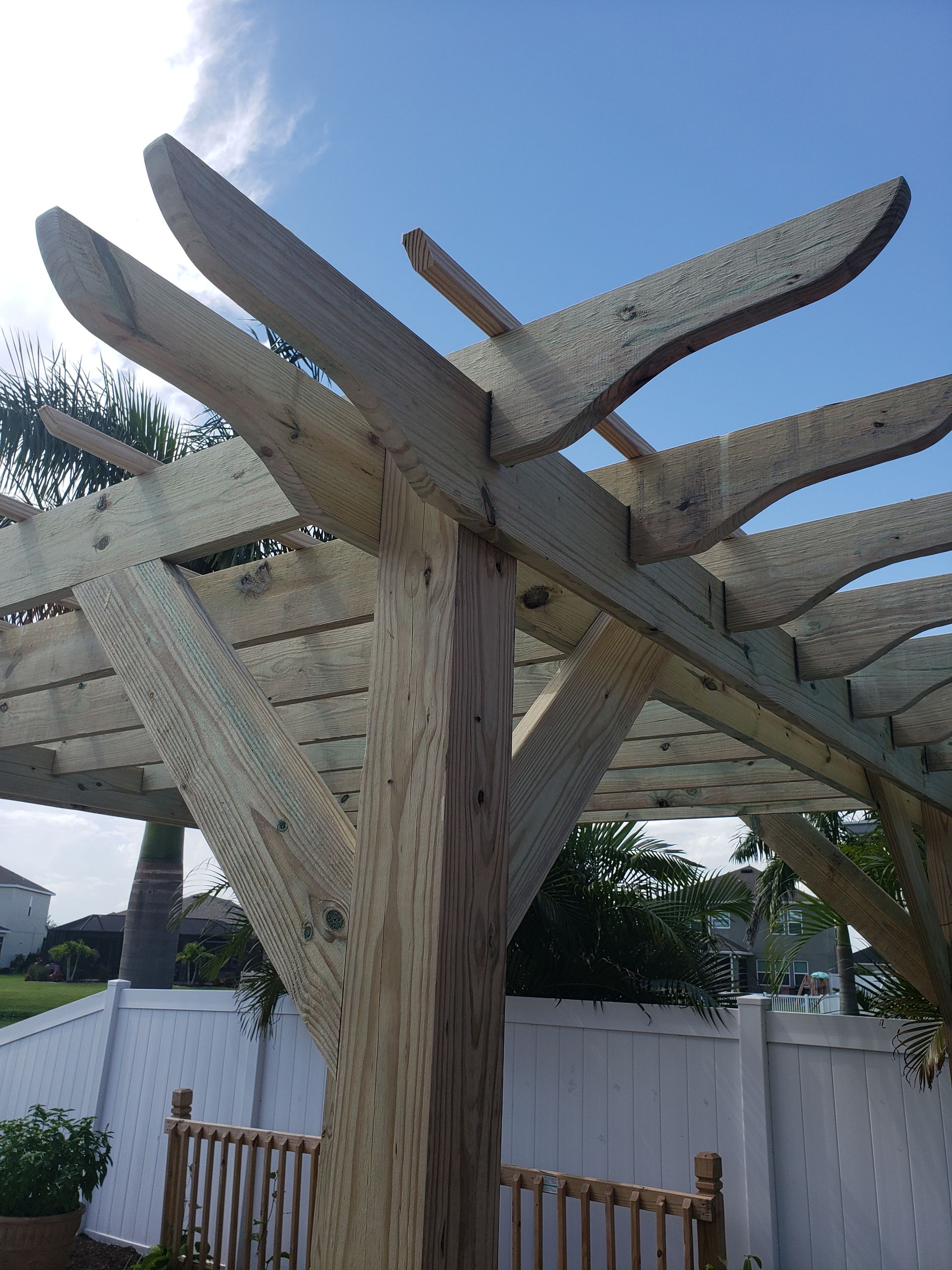 Wooden pergola against a blue sky, with decorative beams and a white fence in the background.