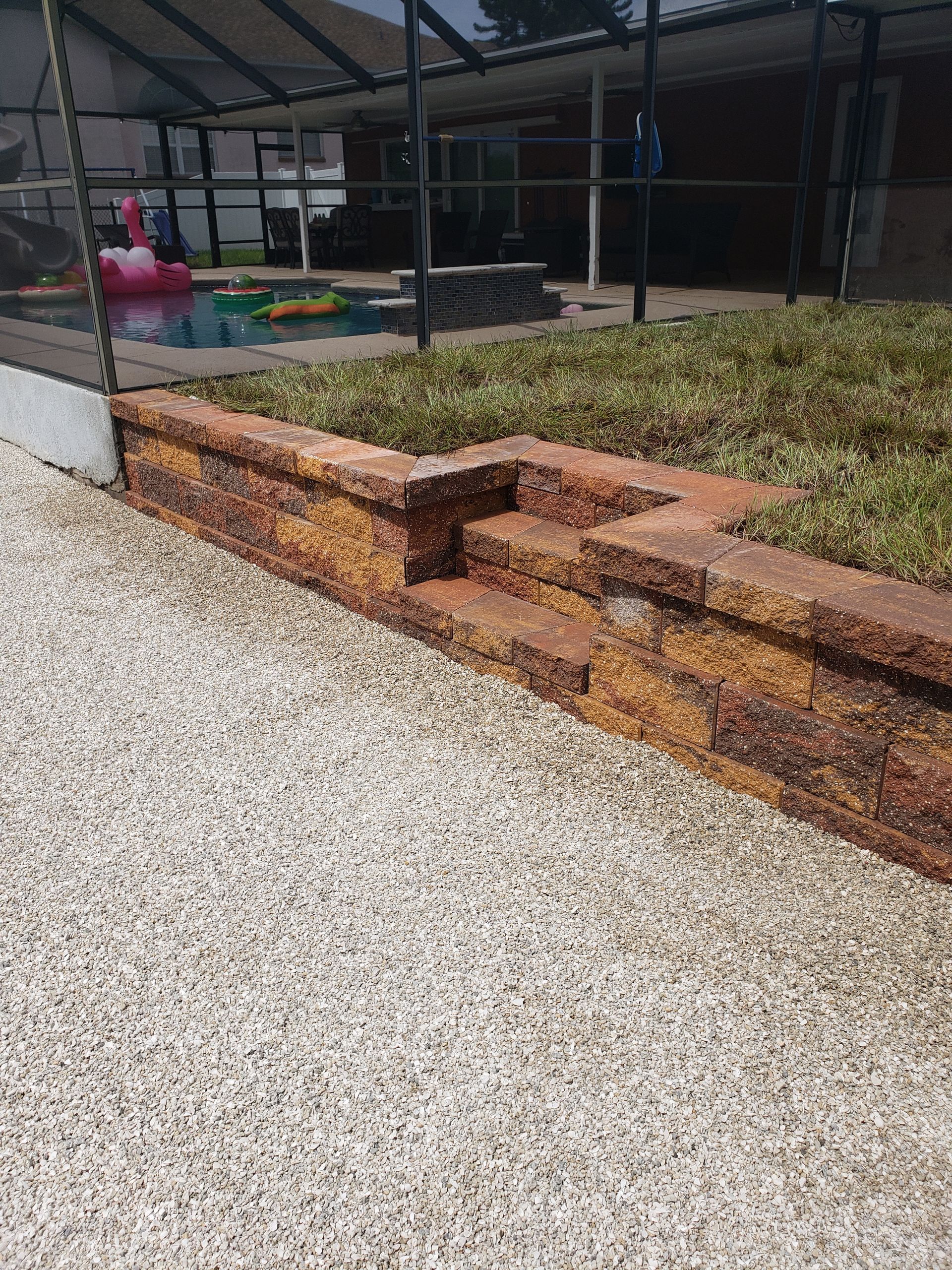 Stone retaining wall with steps beside a gravel area. A pool and grass are visible in the background.