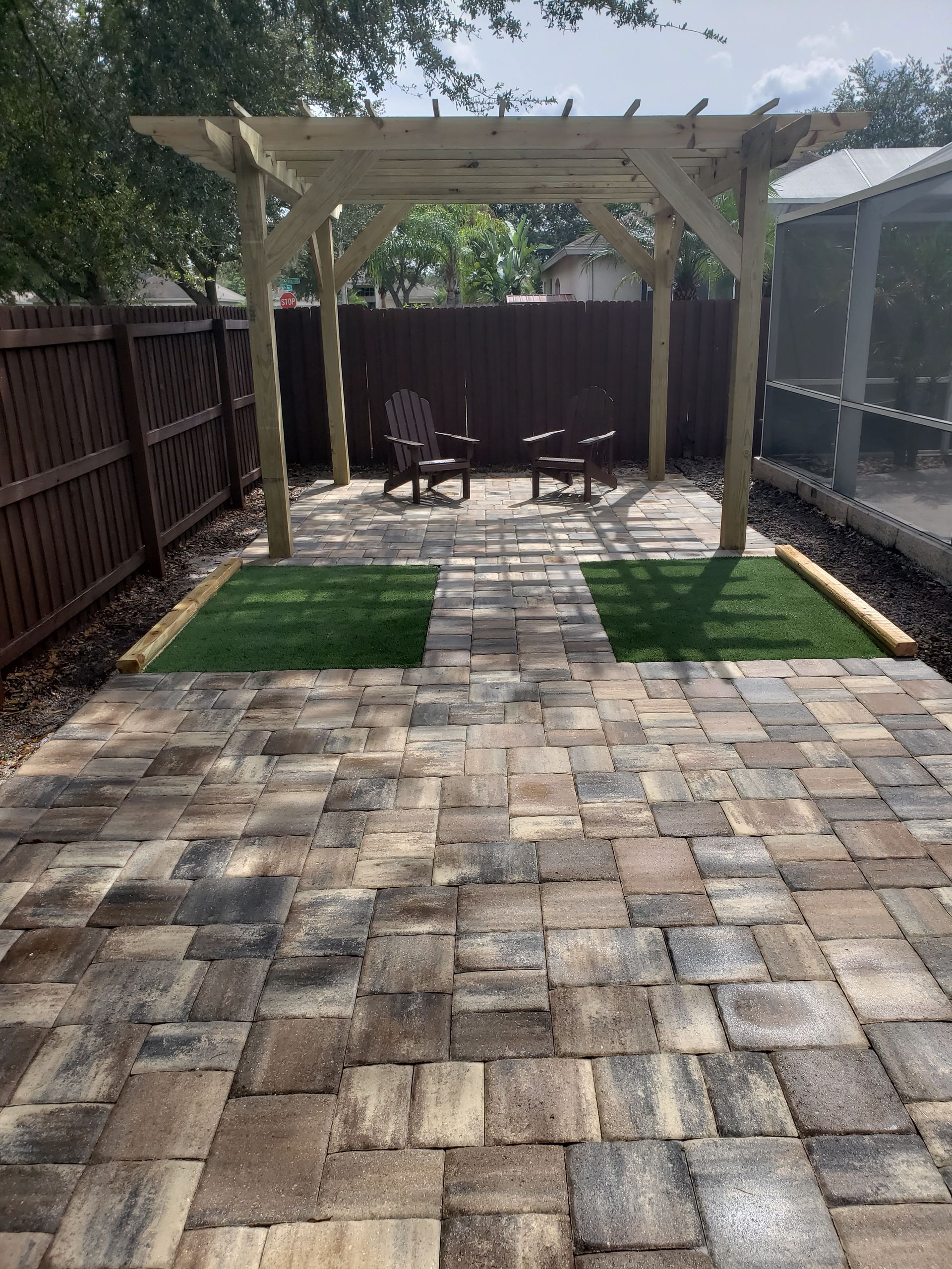 Backyard patio with a pergola, paving stones, artificial grass, and two chairs. Brown fence and a screen enclosure are on either side.