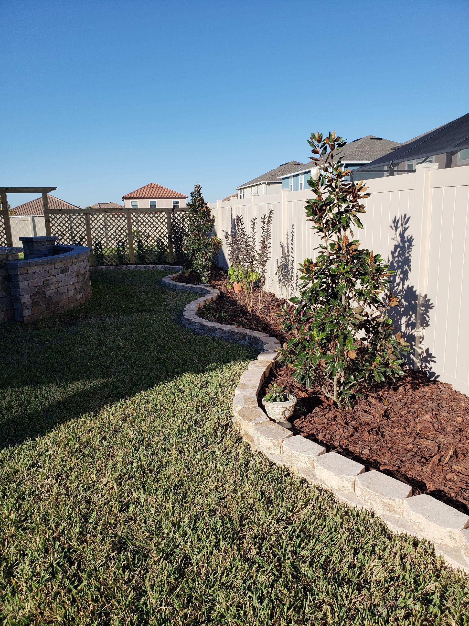 A backyard with a curved stone border around mulch and young trees, along a white fence and green lawn under a blue sky.
