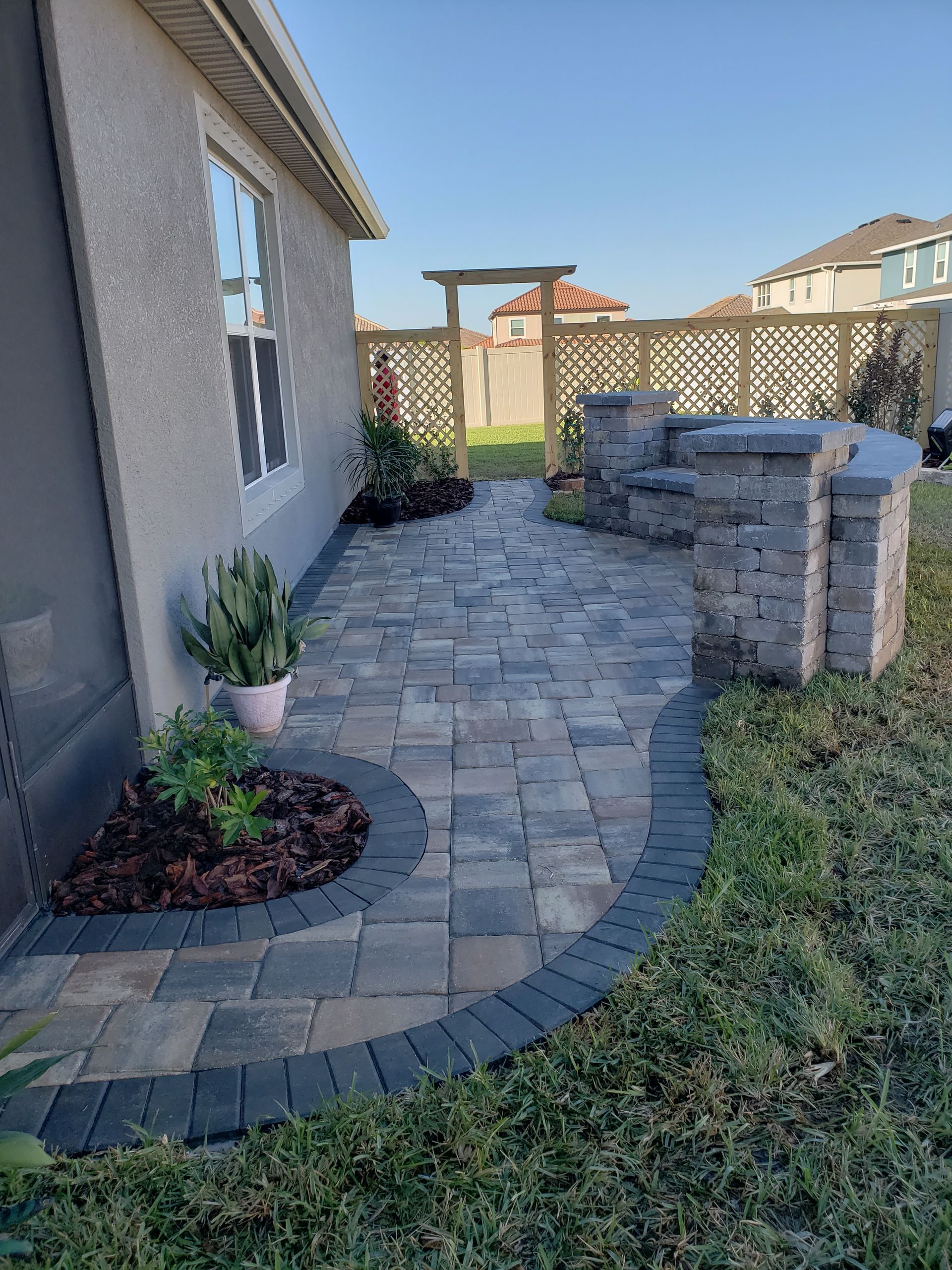A paved patio with a built-in seating area, bordered by grass and a house. A decorative wooden archway stands beyond the patio.