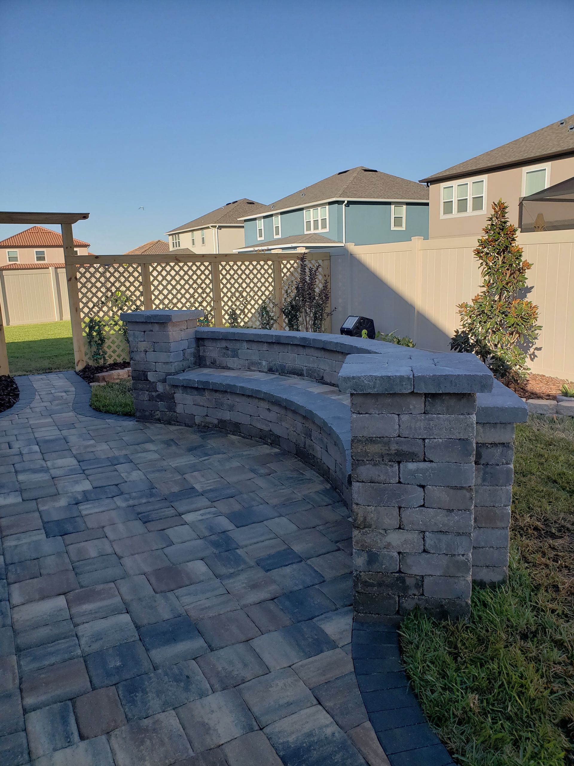 Brick patio with a curved stone bench in a backyard, with a wooden trellis and houses in the background.