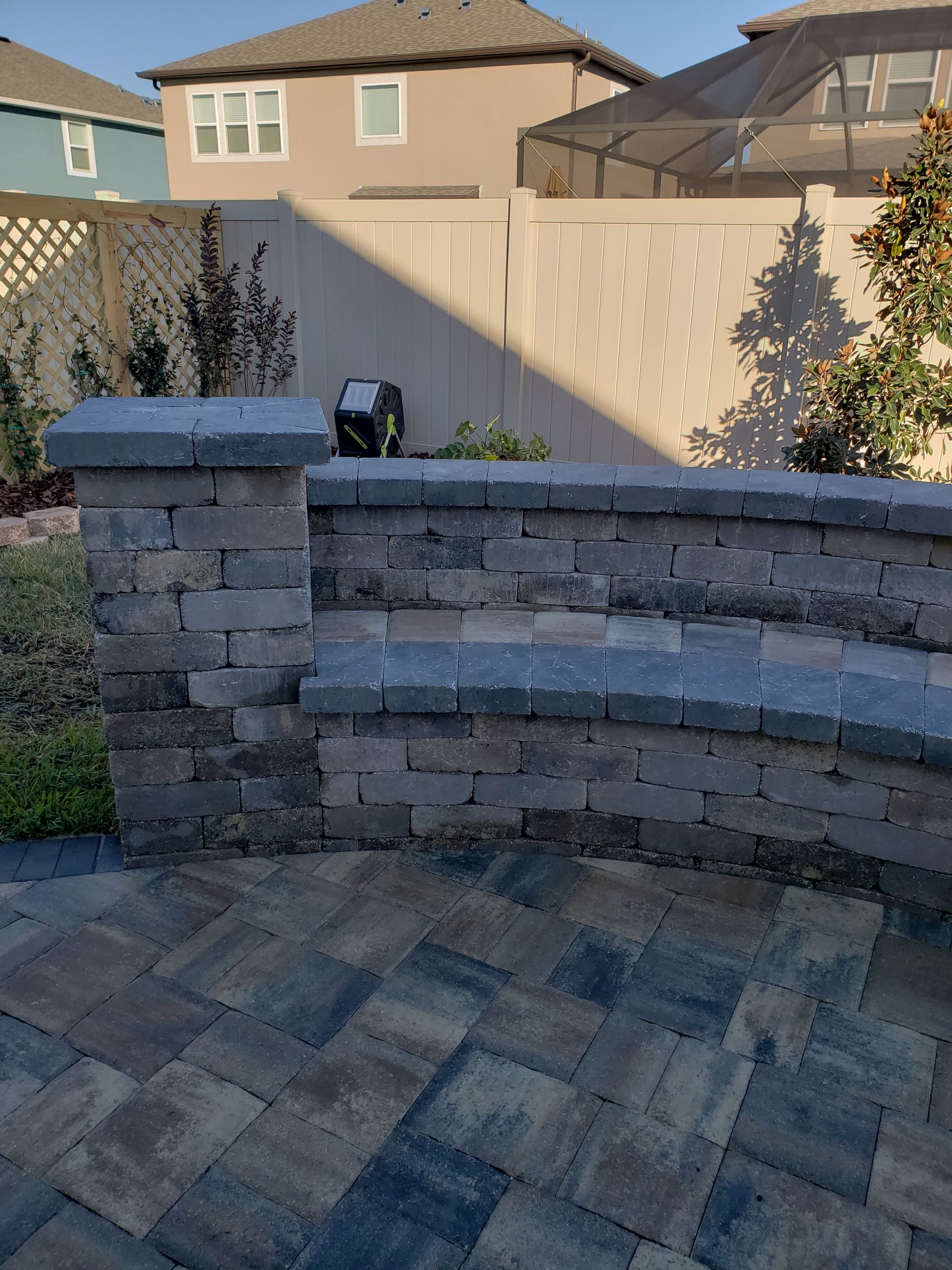 Stone patio with a curved retaining wall and a tall column in a residential backyard.