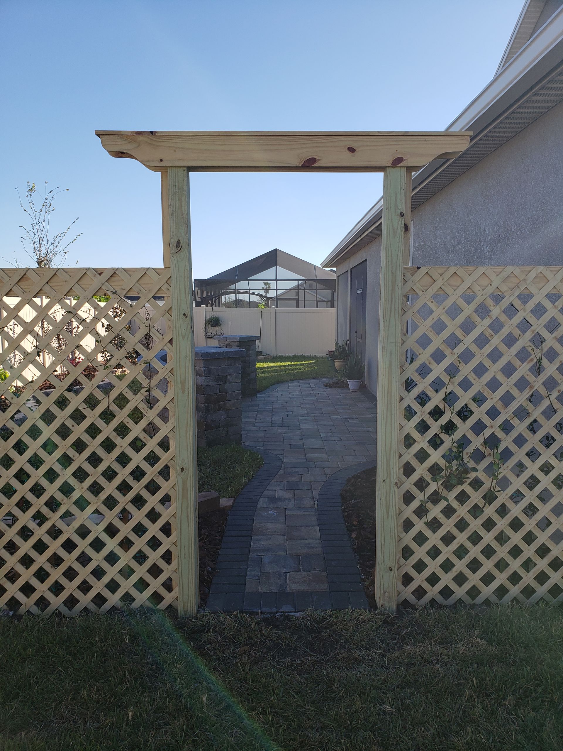 Wooden arbor doorway leading to a brick path and garden, flanked by lattice fencing; bright blue sky in the background.
