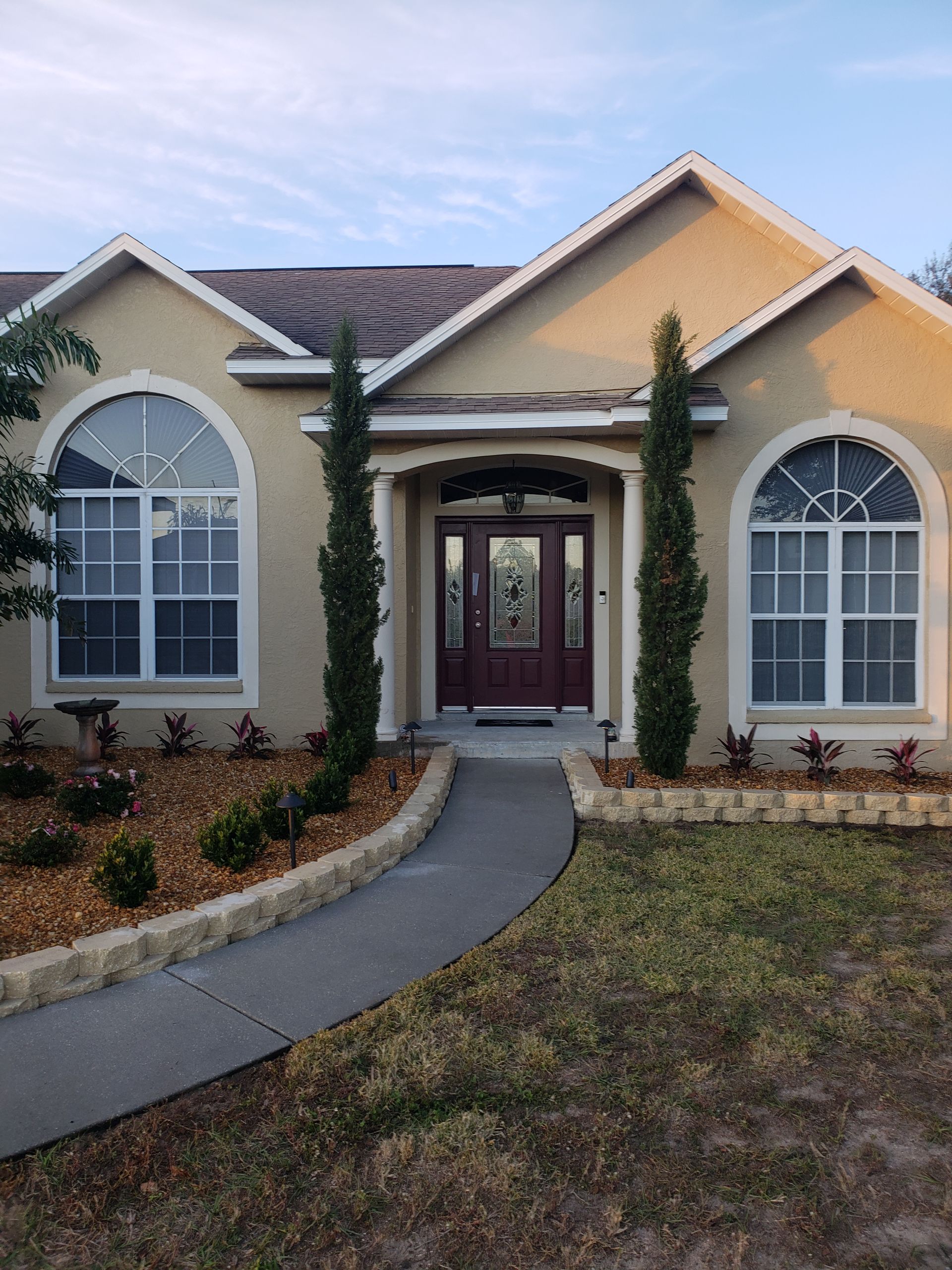 A beige stucco house with arched windows, a burgundy door, and a concrete path leading to the entrance. Two tall, thin trees frame the door.