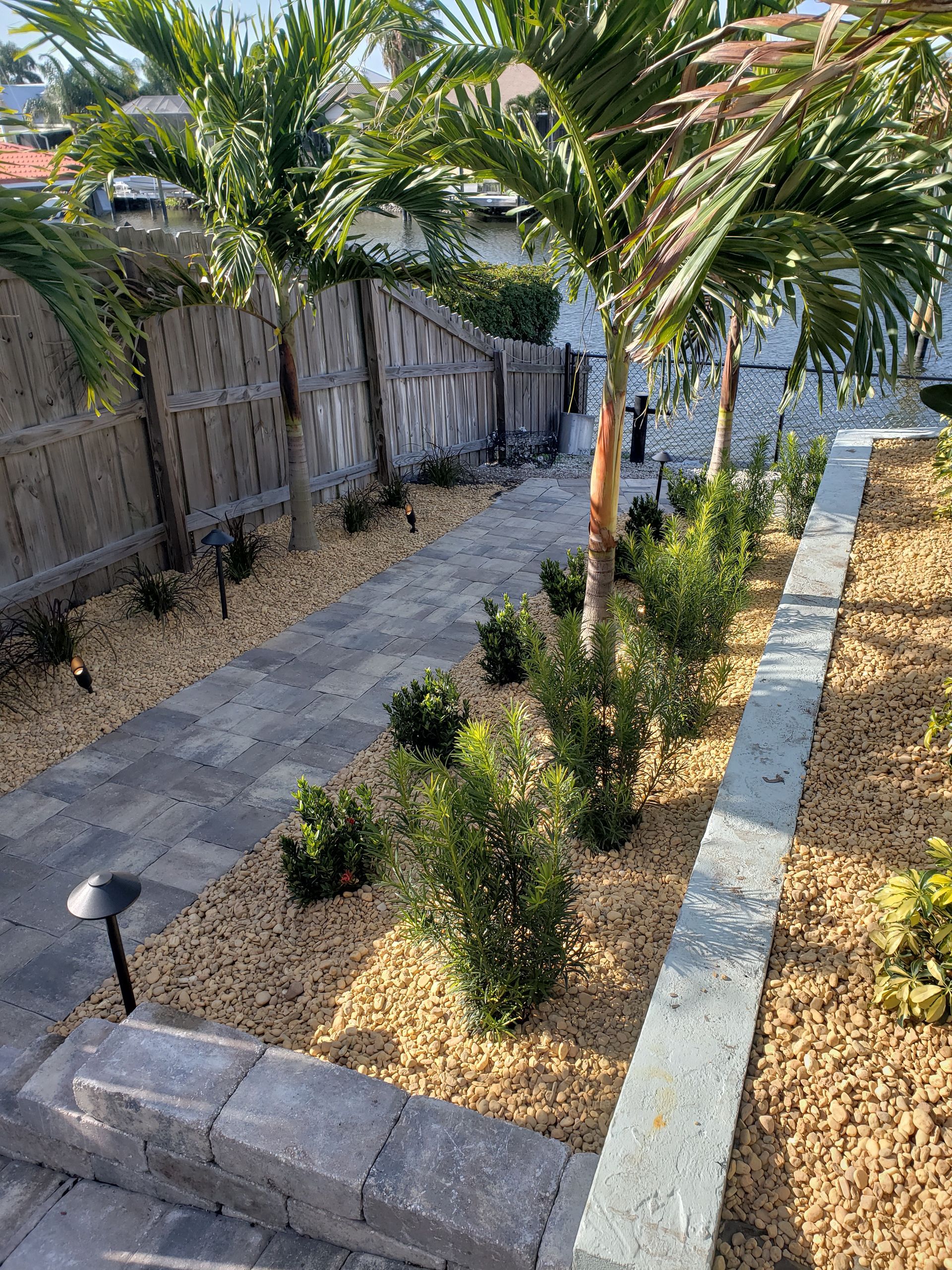 A walkway with pavers and tan gravel, bordered by shrubs and palm trees, alongside a gray retaining wall.