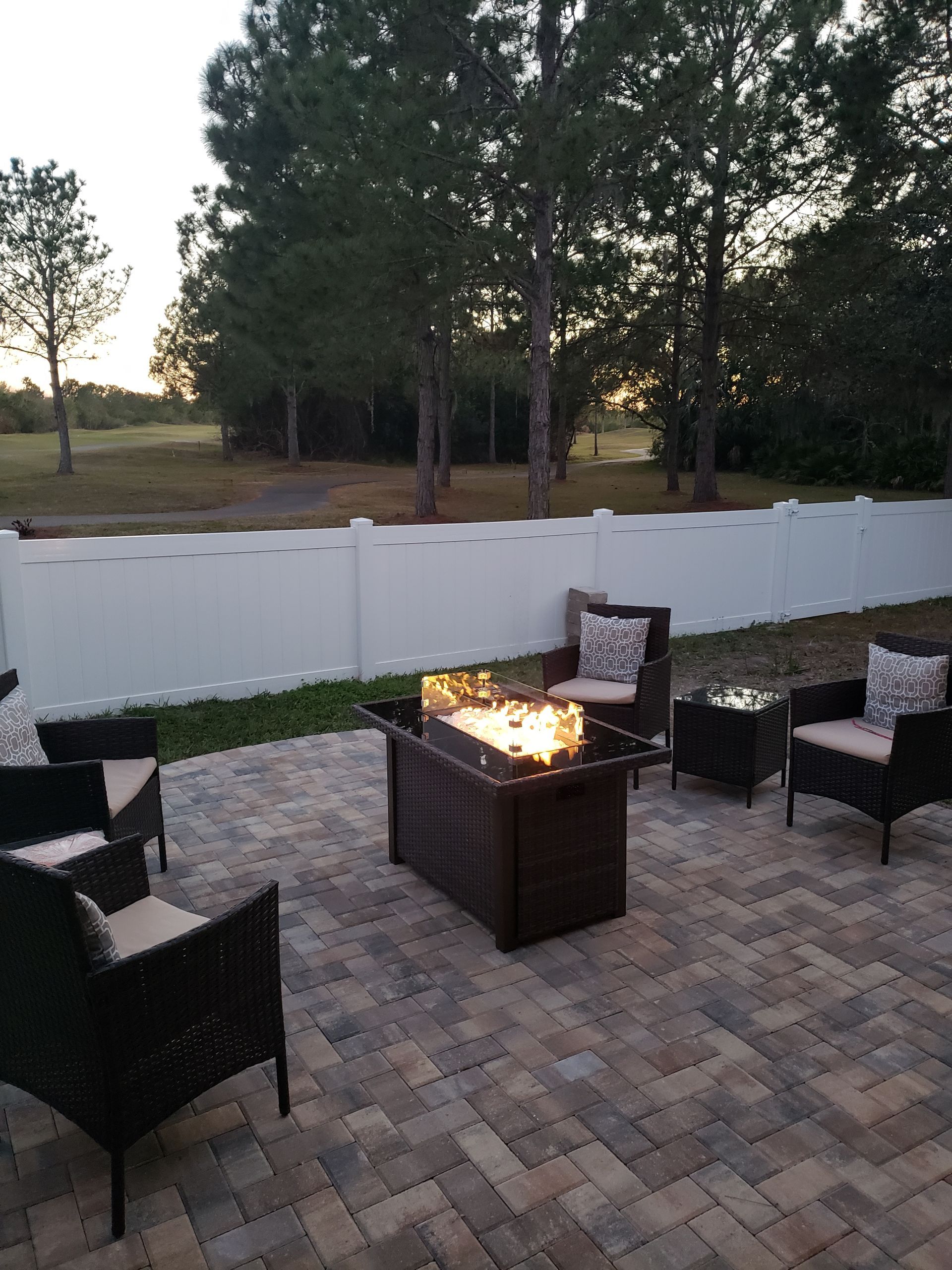 Outdoor patio with fire pit surrounded by wicker chairs, bordered by a white fence and trees.