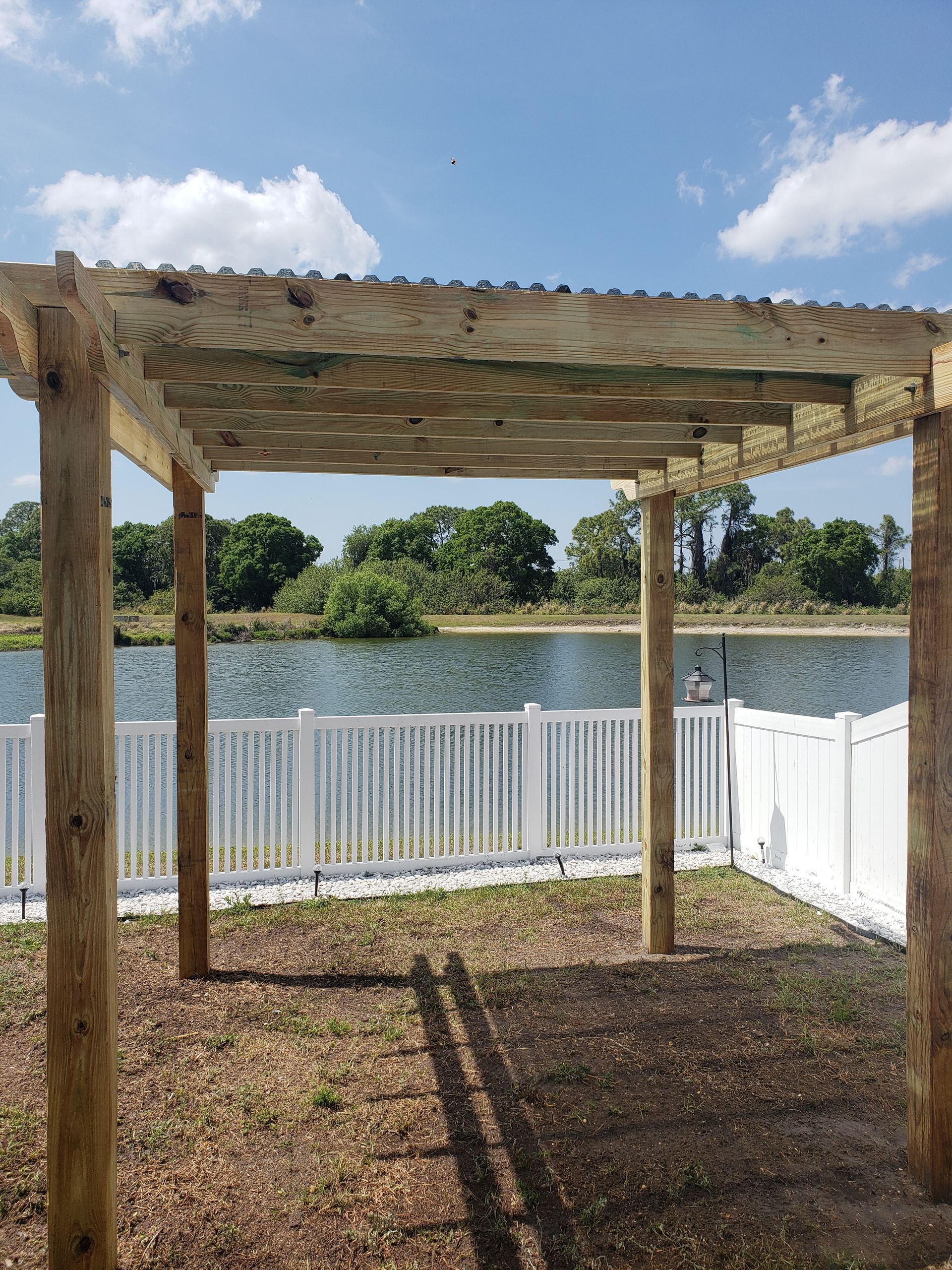 Wooden pergola overlooking a lake, white fence, and blue sky. Sunlight casts shadows on the ground.