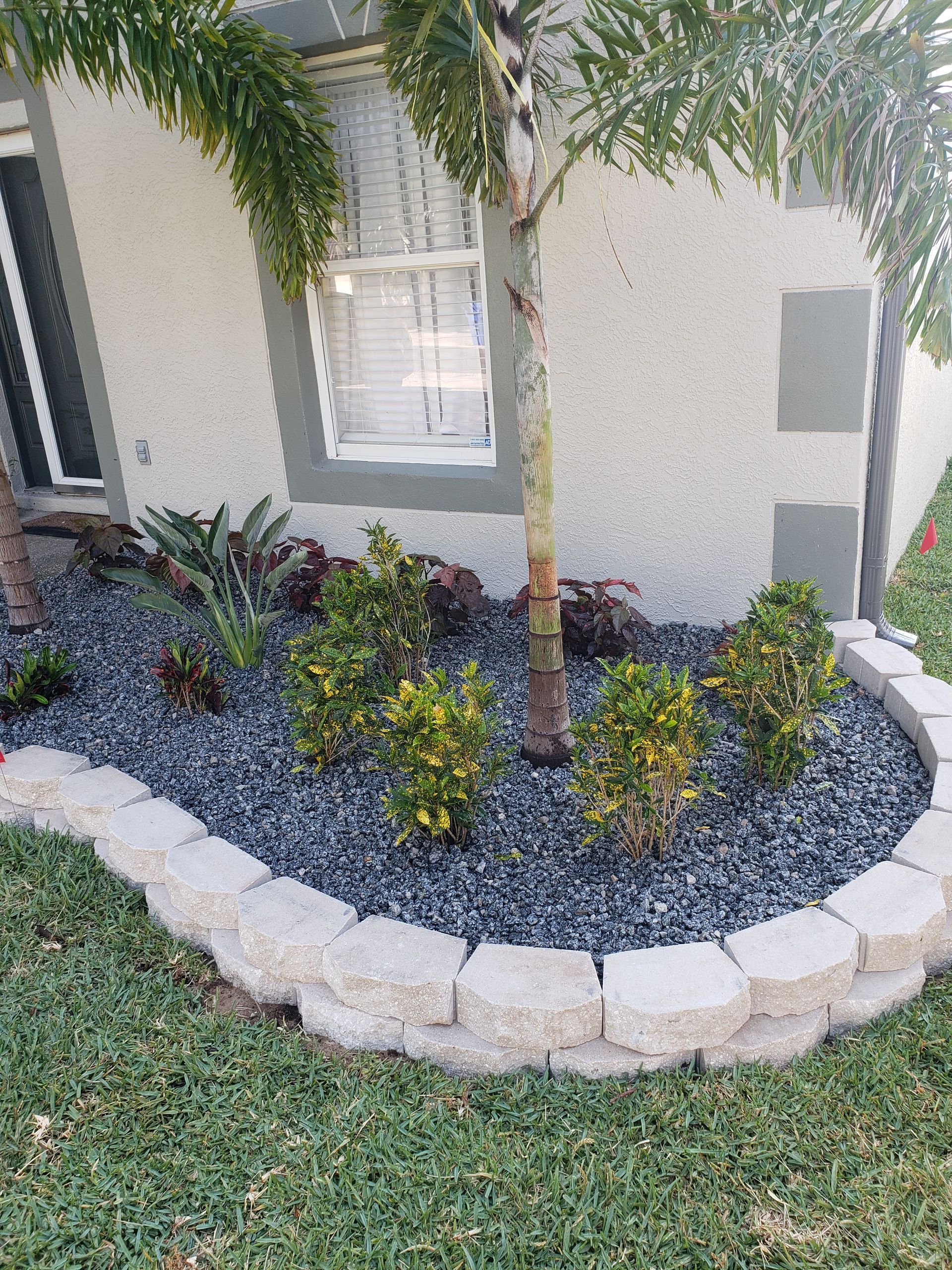 A landscaped garden bed with dark gray pebbles, bordered by light-colored bricks, with green plants and a palm tree against a stucco wall.