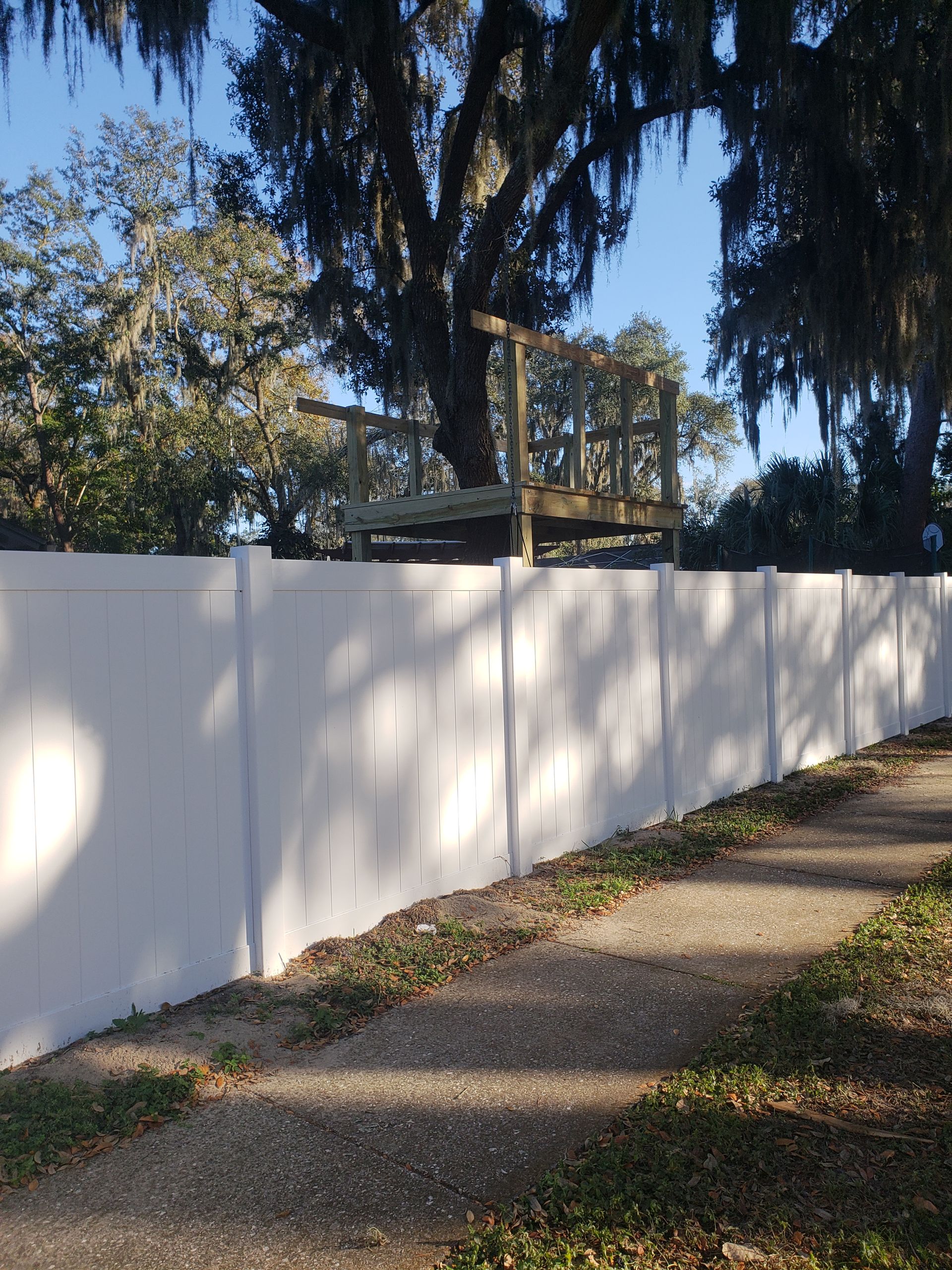 White fence in front of a small wooden deck built around a tree, beside a gravel path and grass.