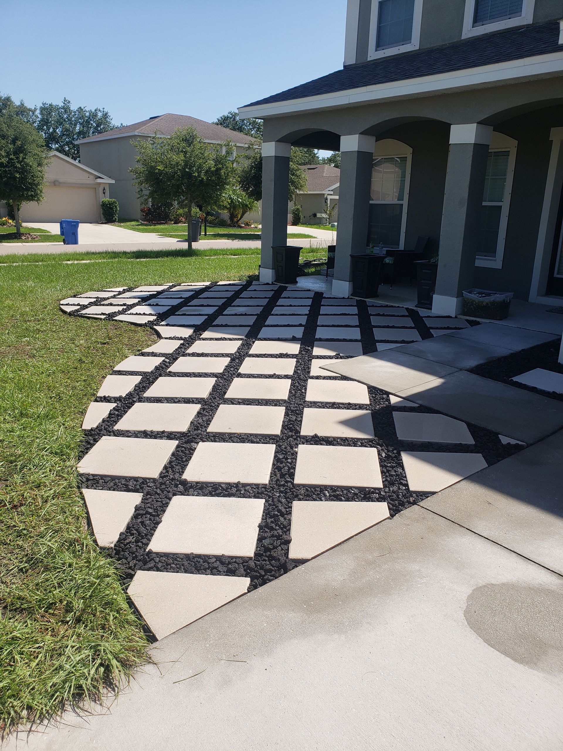 Stone patio pathway with square pavers and black gravel leading to a house with a porch and a green lawn.