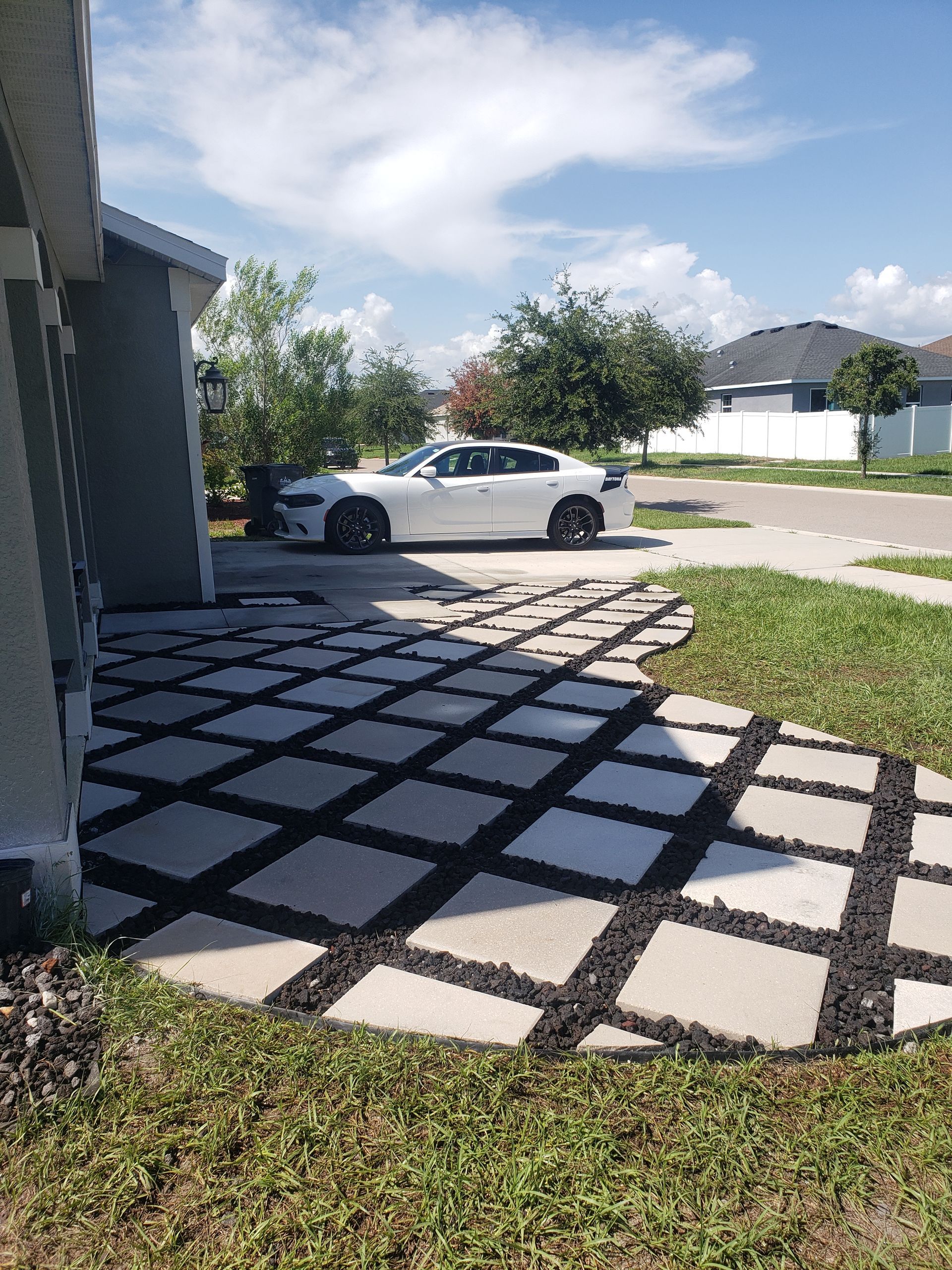 Concrete pavers with black gravel create a walkway near a home's driveway where a white car is parked, under a sunny sky.
