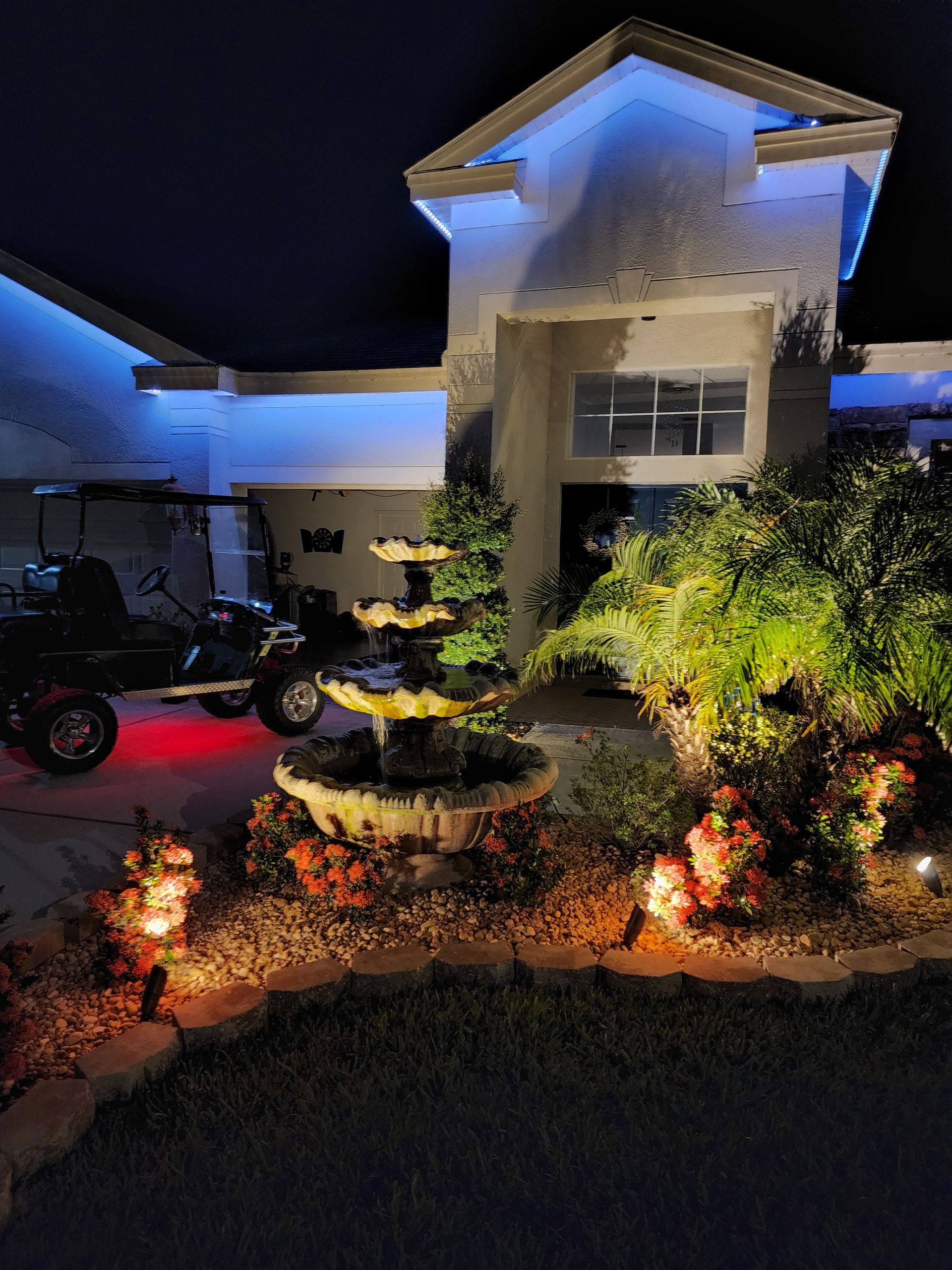 House exterior illuminated at night with blue and orange lights, a fountain, and golf cart. Landscaping with lit plants adds to the ambiance.