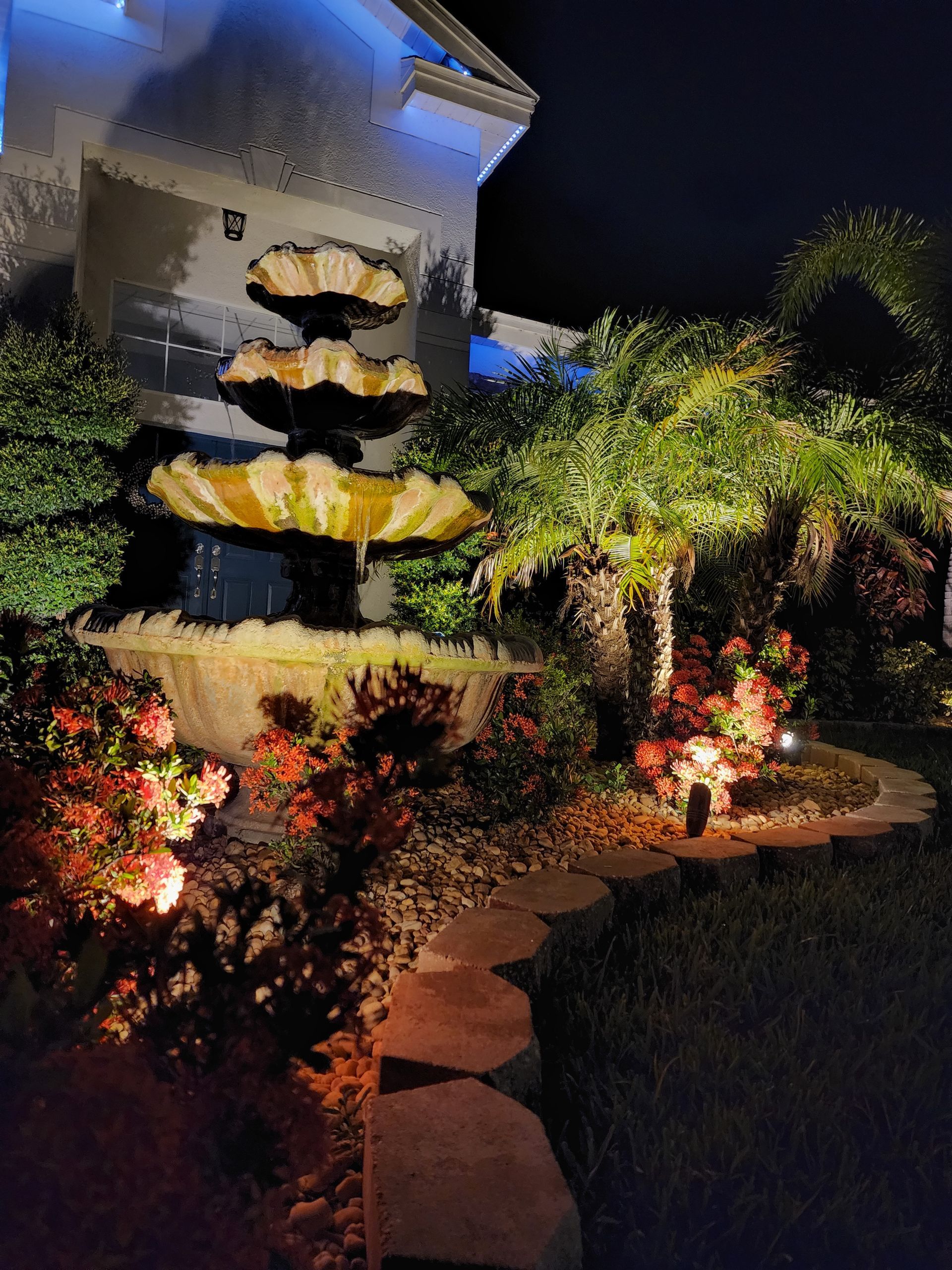 Nighttime view of a lit-up tiered fountain surrounded by illuminated plants and a stone path.