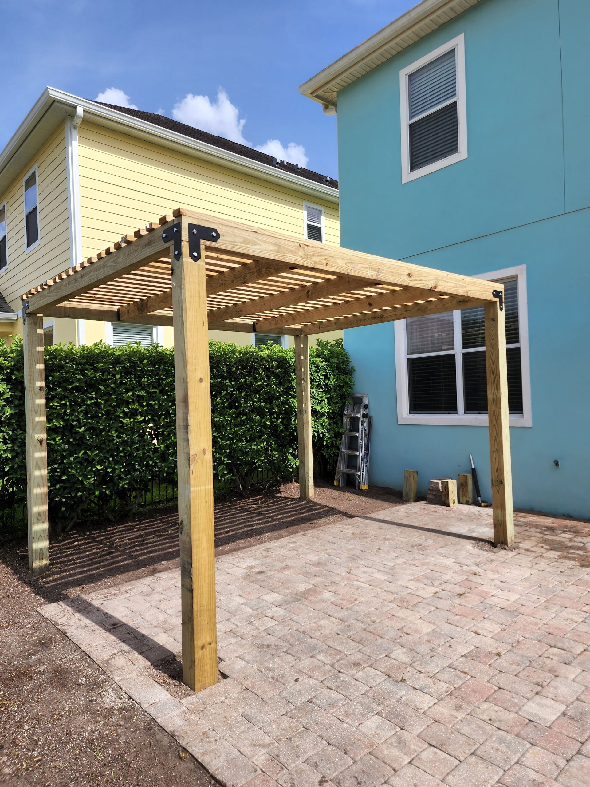 Wooden pergola on a brick patio, flanked by a green hedge and two-story buildings with light blue and yellow facades.