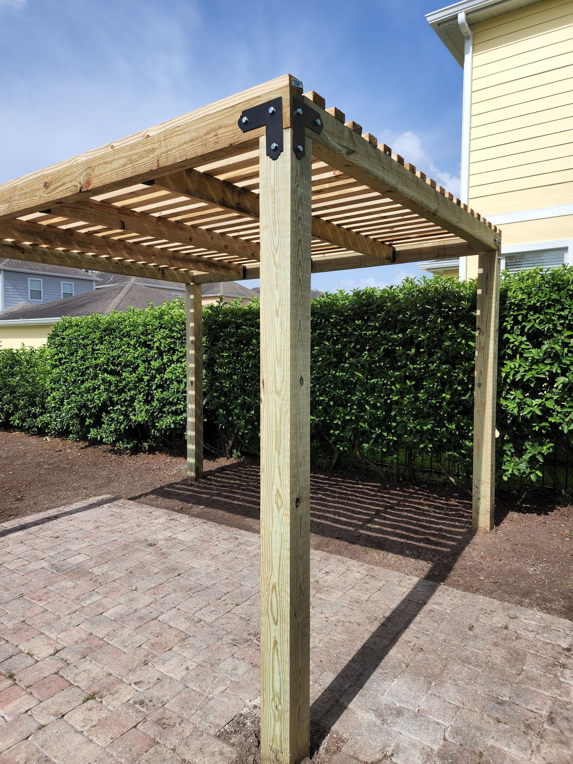 Wooden pergola with a slatted roof casts shadows on a brick patio. Green hedges and a yellow building are in the background.